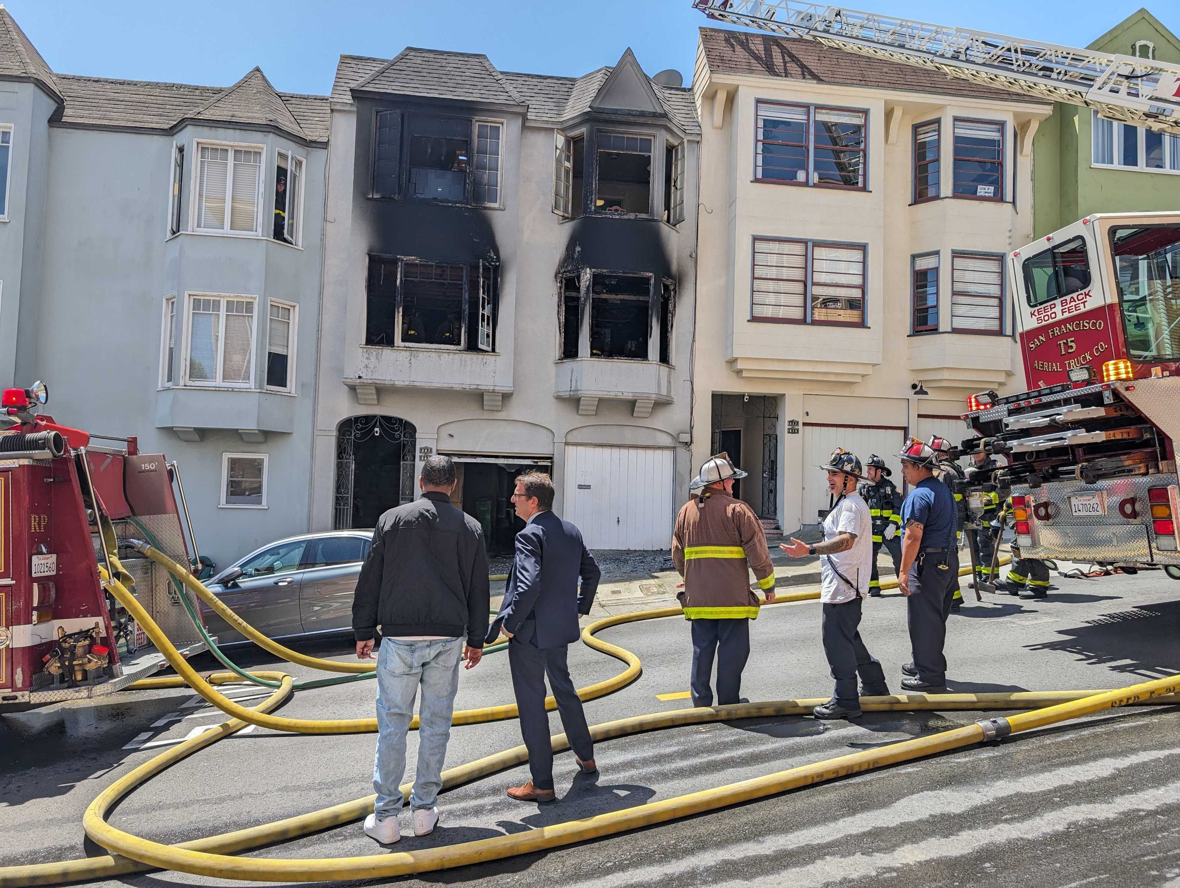 Firefighters work at a fire-damaged building with charred windows, as men overlook the scene, and fire trucks line the street.