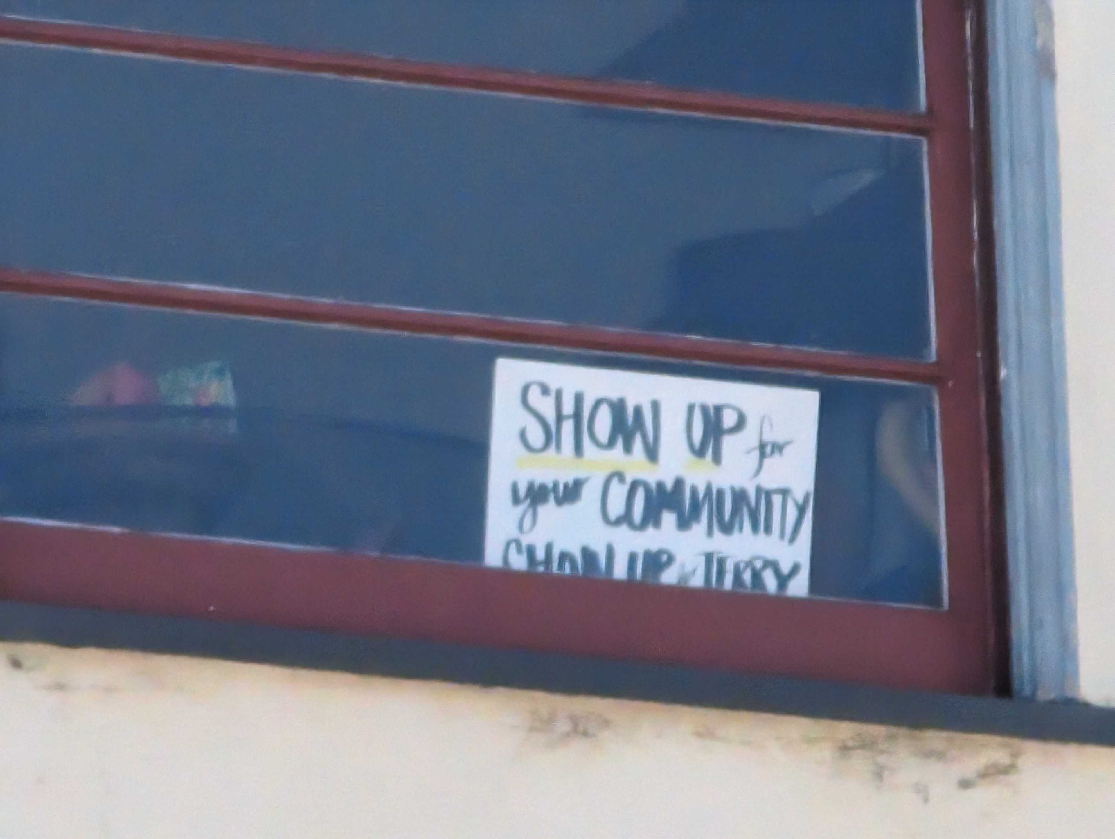 A sign in a window reads &quot;SHOW UP for your COMMUNITY Show up. Vote.&quot; It is handwritten with blue borders.