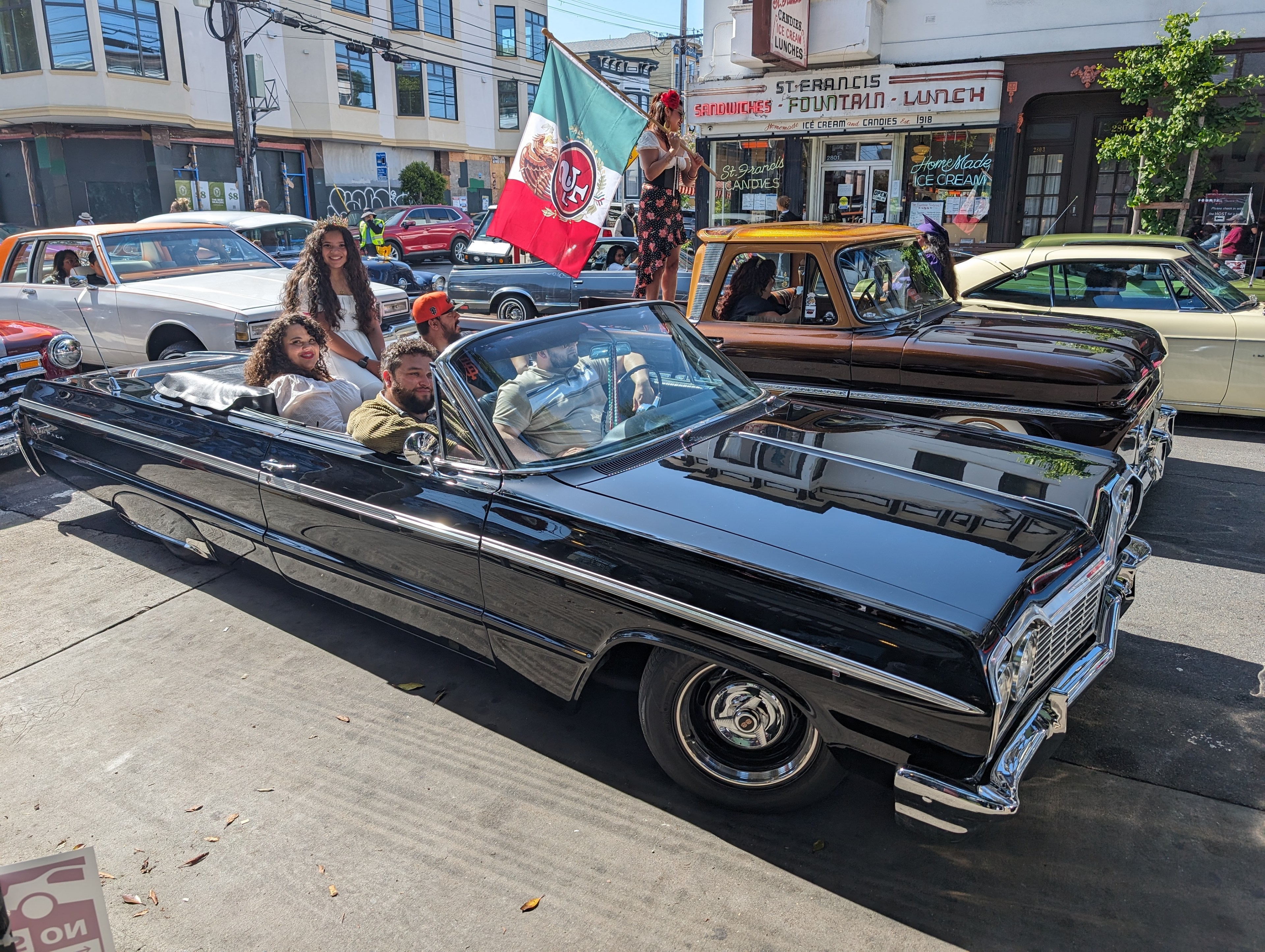 A black Cadillac with five smiling, happy people enjoying bright sunshine before a parade starts.