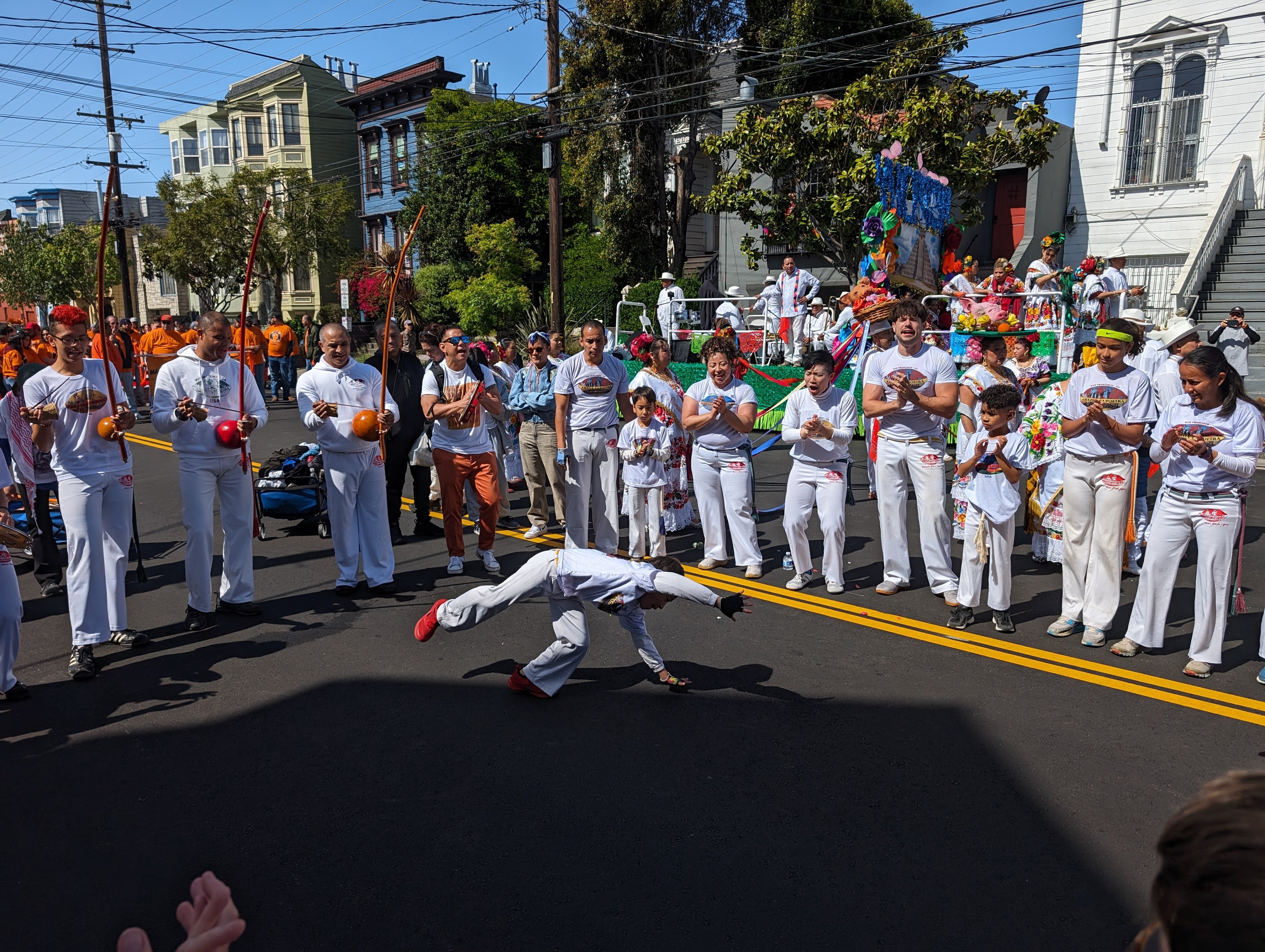 A white-clad boy wearing red shoes cartwheels in a street as white-clad dancers and musicians play around him in a circle.