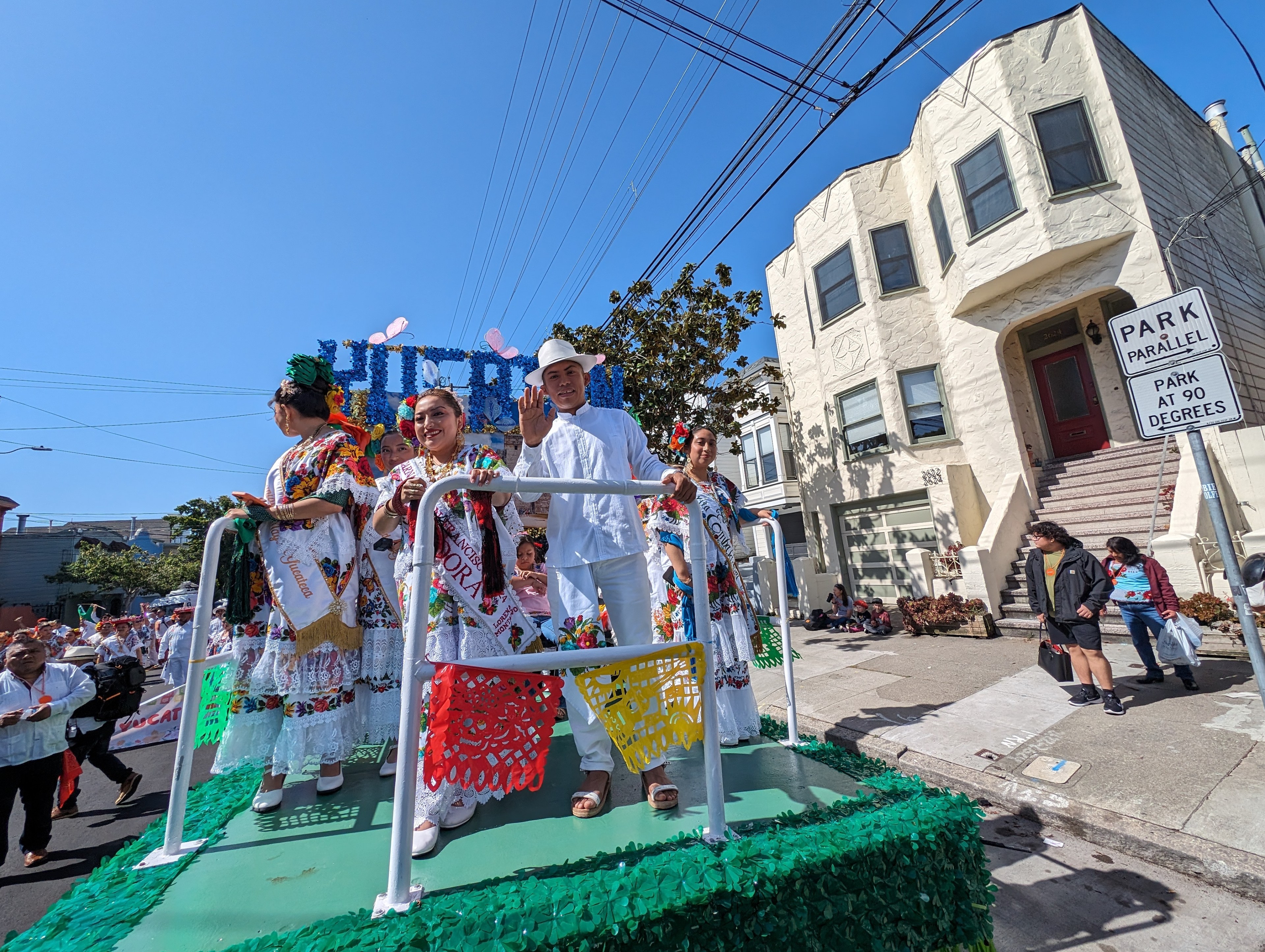 A green float with white railings waits along a city street curb as several people wearing ornate and colorful outfits smile and wave at a camera.