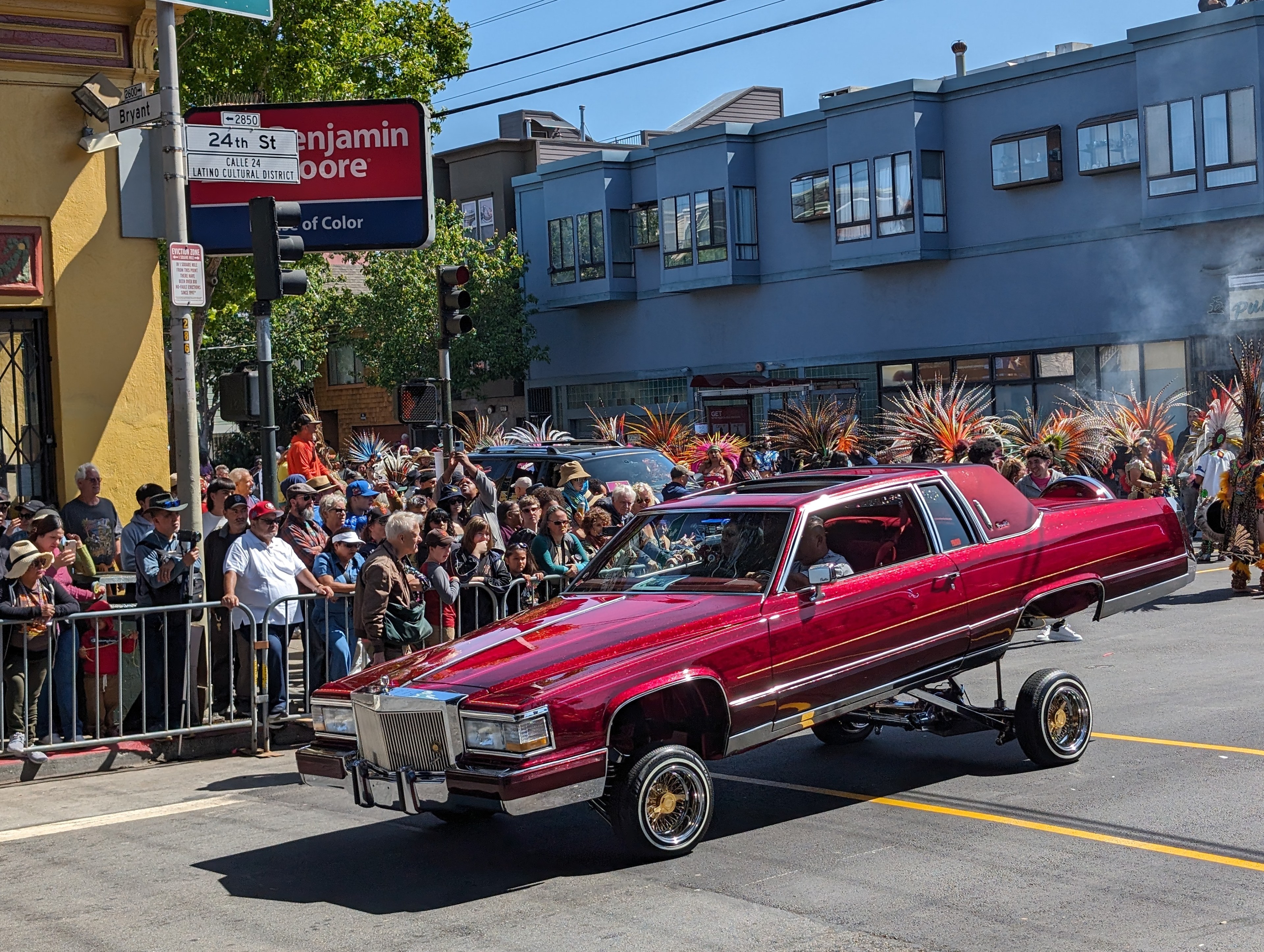 A candy apple red Cadillac on lifted wheels rides through a city intersection.