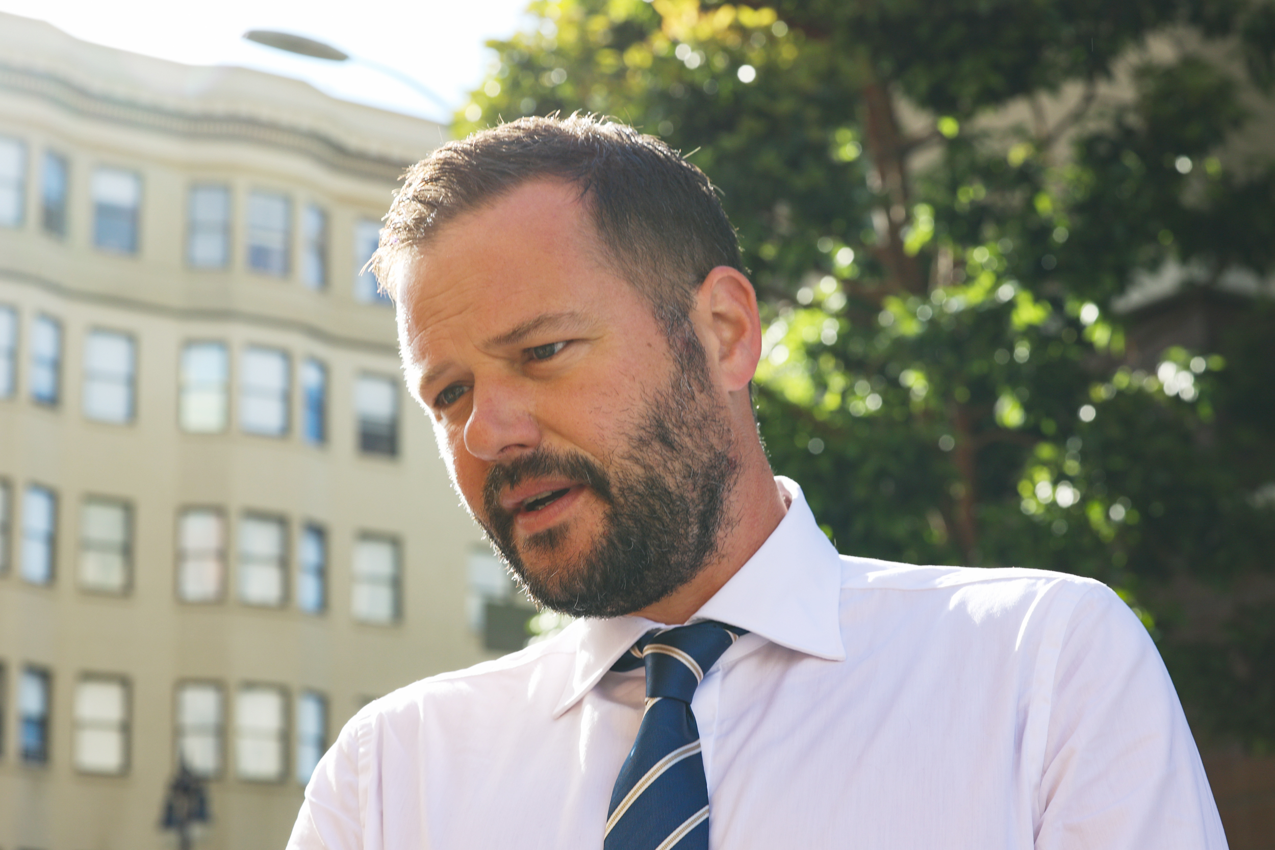 A man with a beard wearing a white shirt and blue tie is pictured outdoors. Behind him are blurred trees and apartment buildings in sunlight.