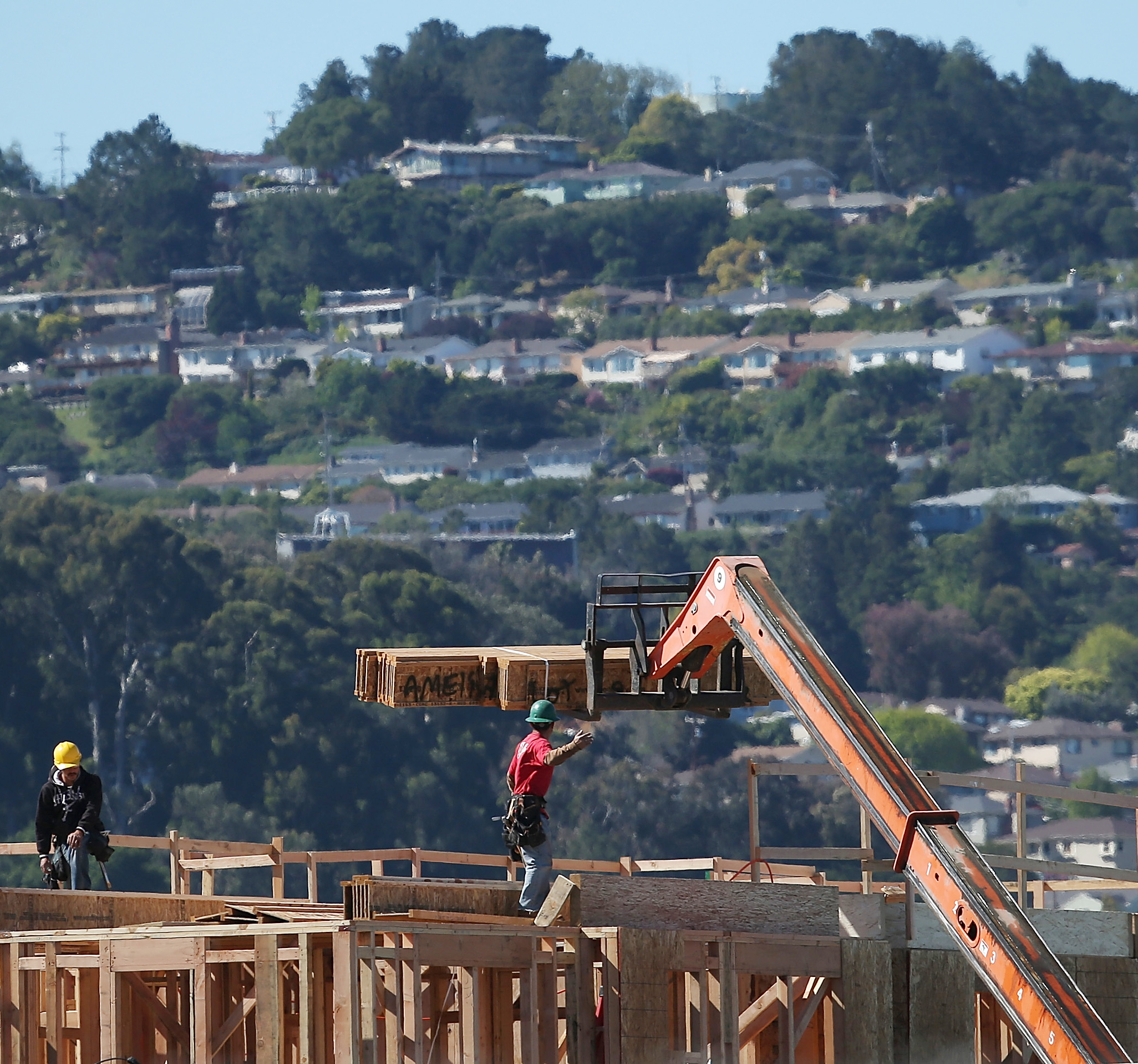 Two construction workers in helmets are on a wooden building frame. One directs a crane lifting a beam, while a hilly residential area is visible in the background.