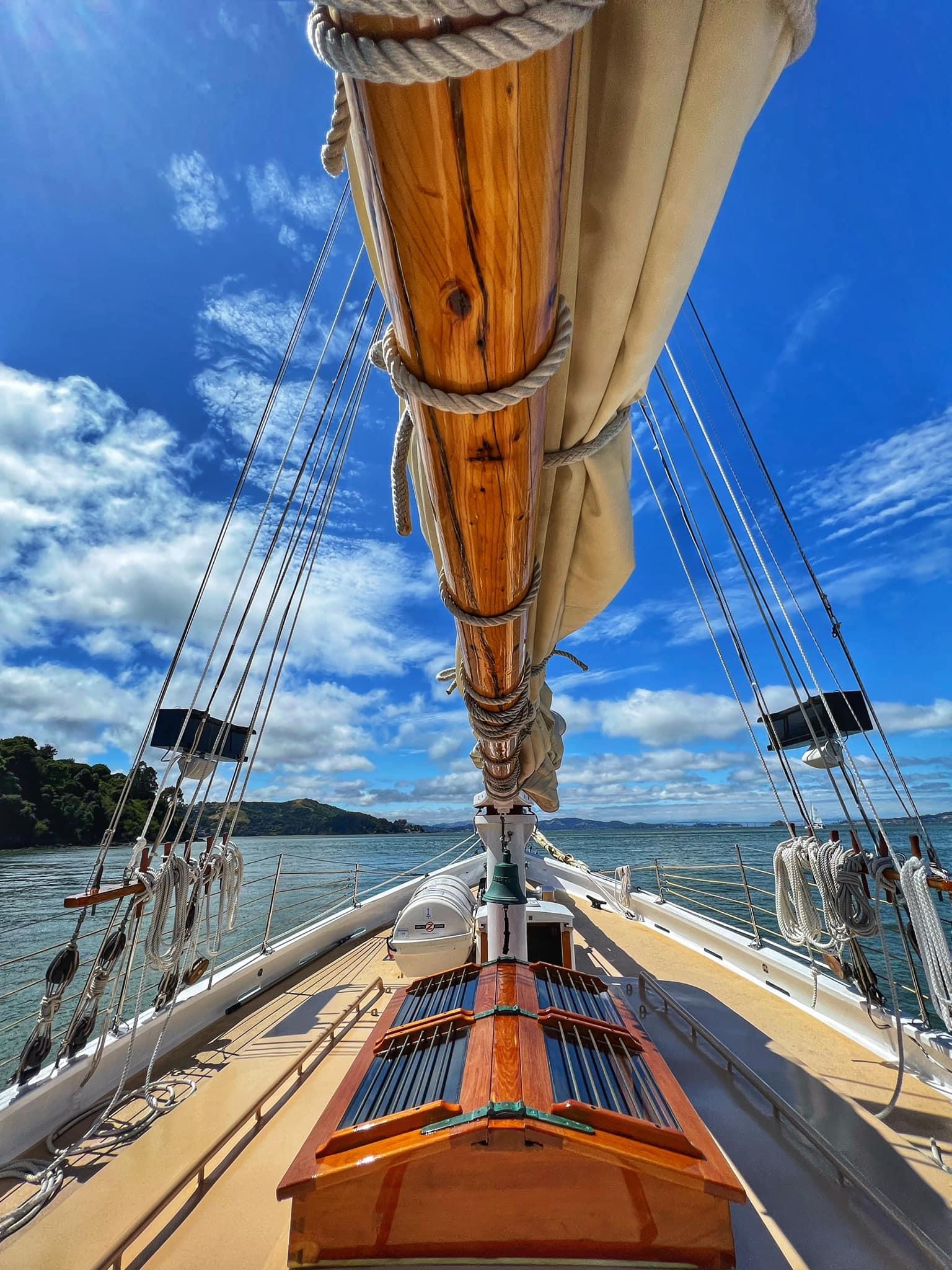 A sailboat's bow with a furled sail, polished wood, riggings, and a blue sky with clouds above.