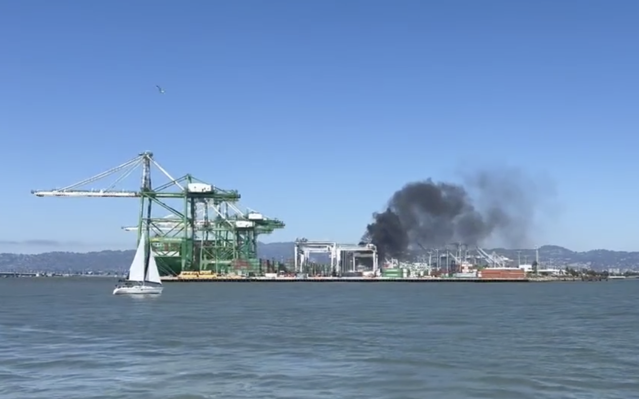 A sailboat glides on water near a dock with green cranes; smoke rises in the background from a distant fire.