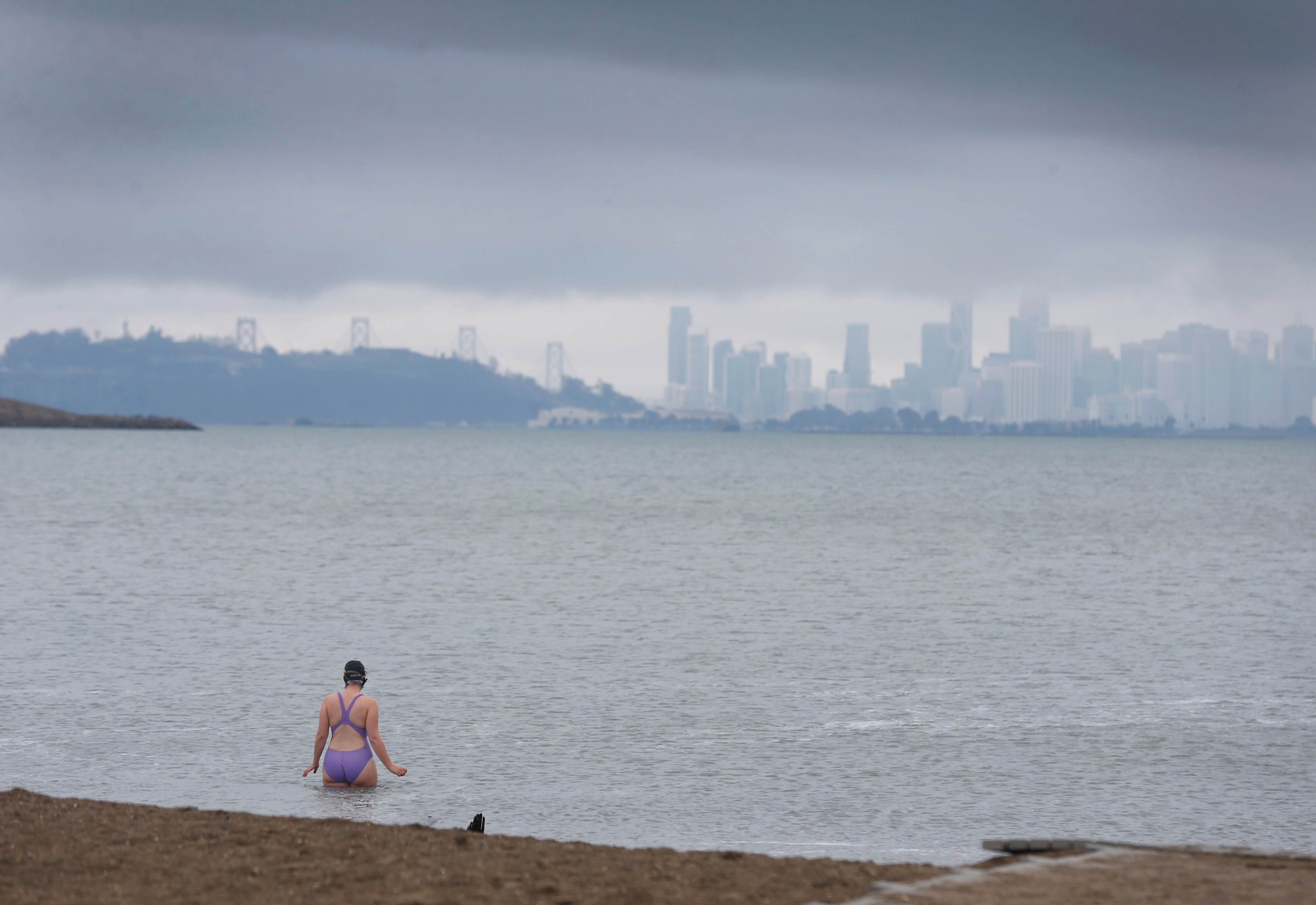 A person in a swimsuit sits by the water, looking at a foggy city skyline in the distance.