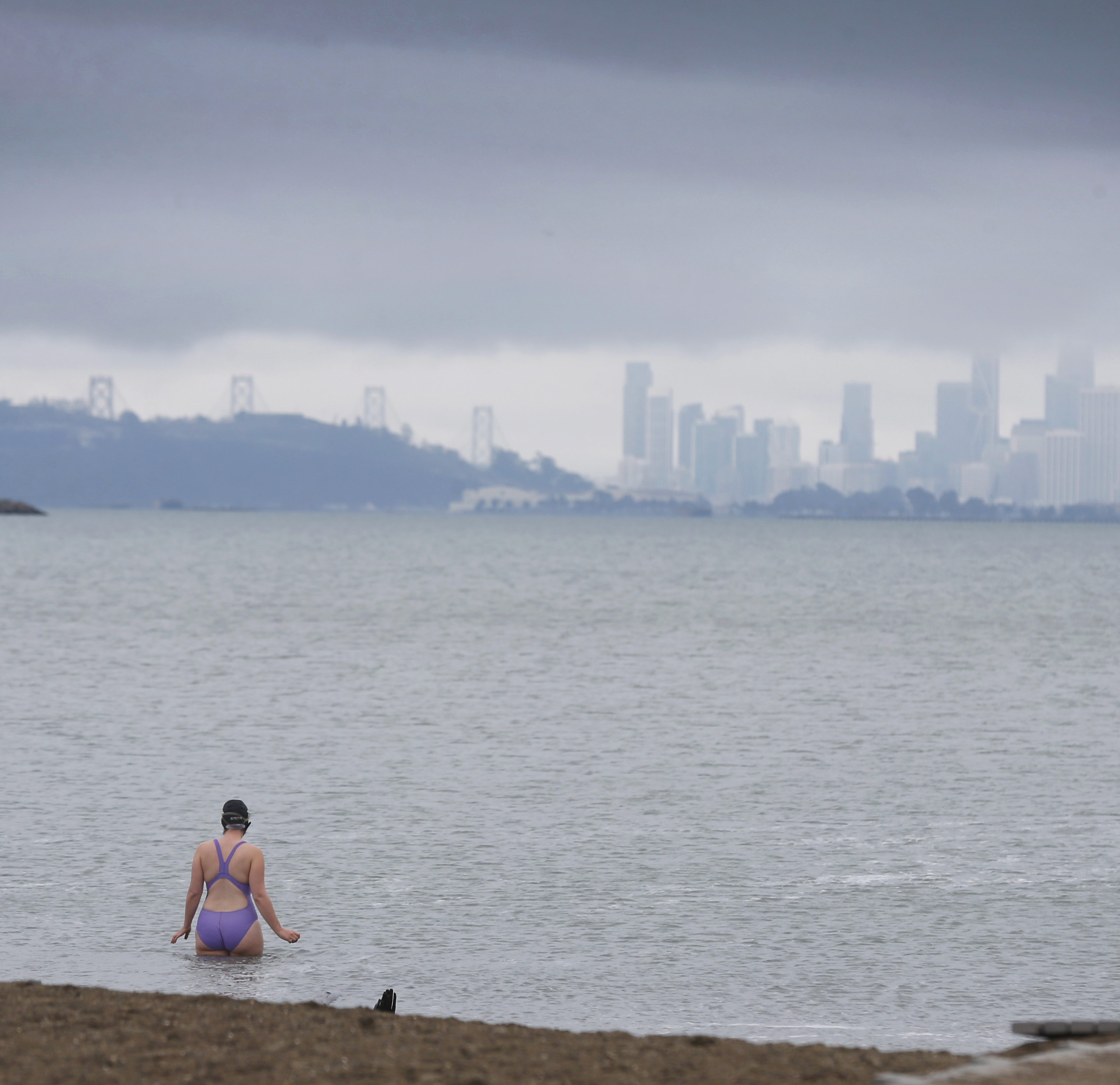 A person in a swimsuit sits by the sea; a foggy city skyline with a bridge lurks in the background.