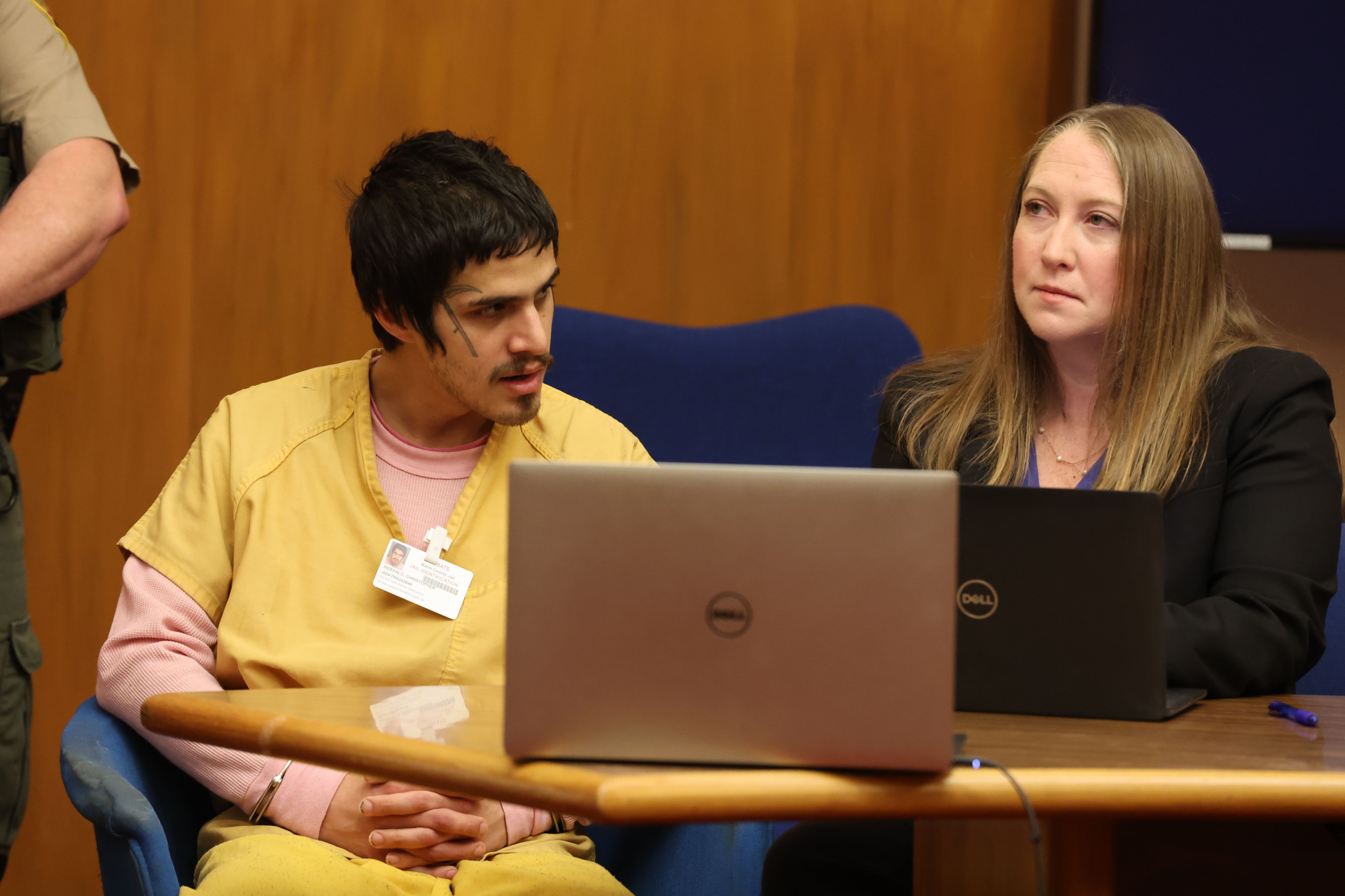 A man in a yellow outfit sits by a woman with a laptop, both at a wooden table, possibly in a courtroom setting.