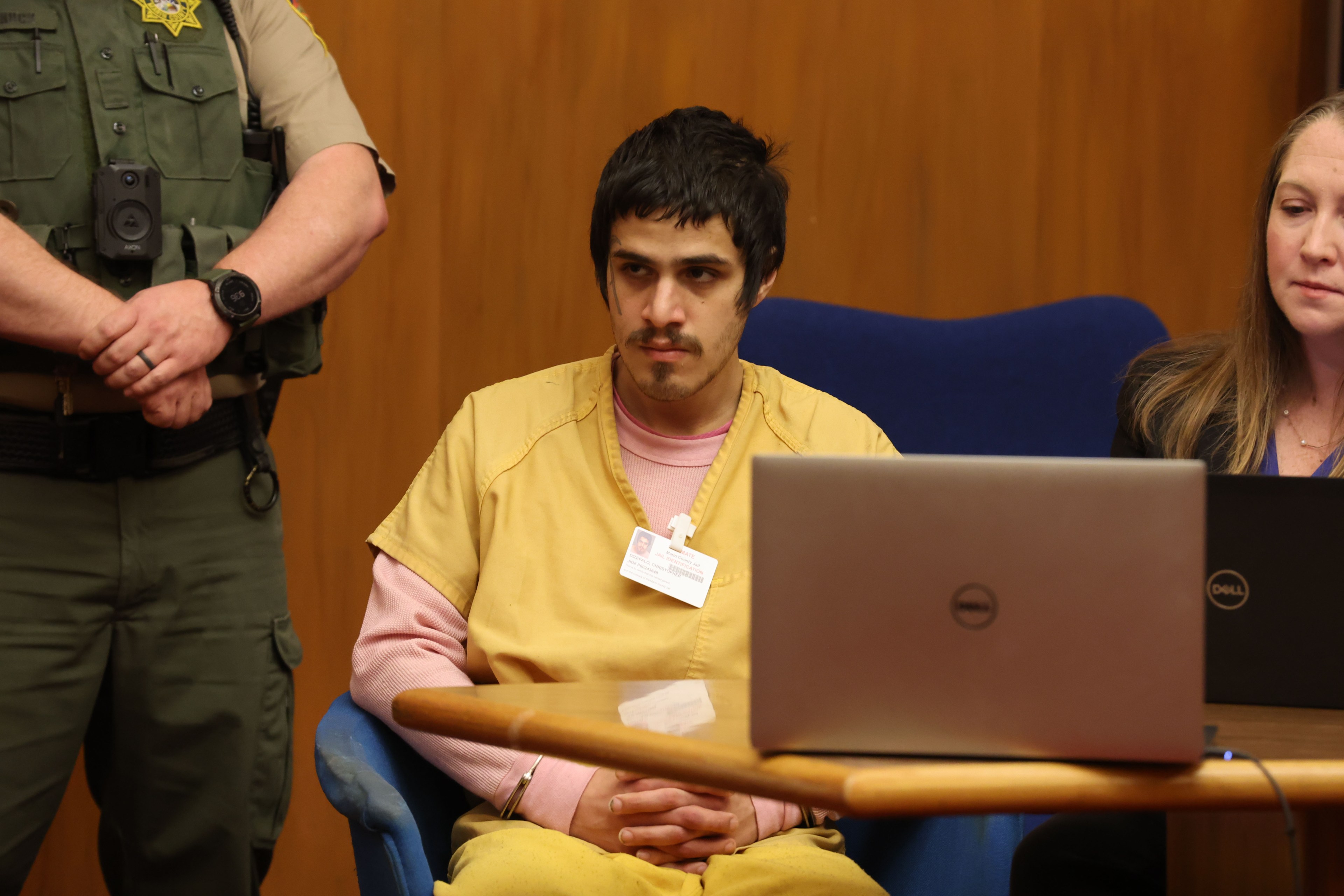 A person in a yellow jumpsuit sits by a laptop, flanked by an officer and another individual.