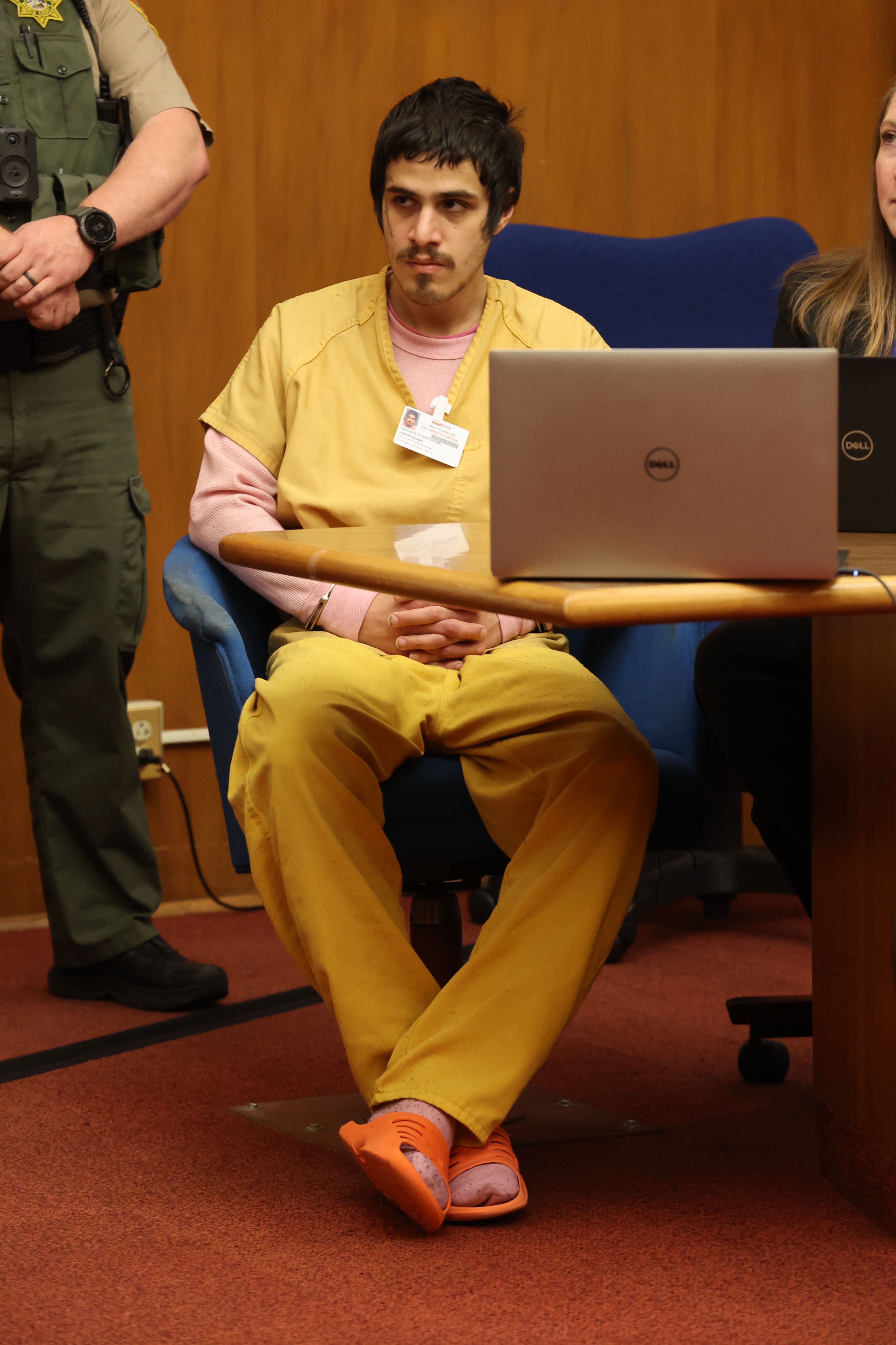 A man in a yellow prison uniform sits by a laptop, next to a woman and partially shown officer.