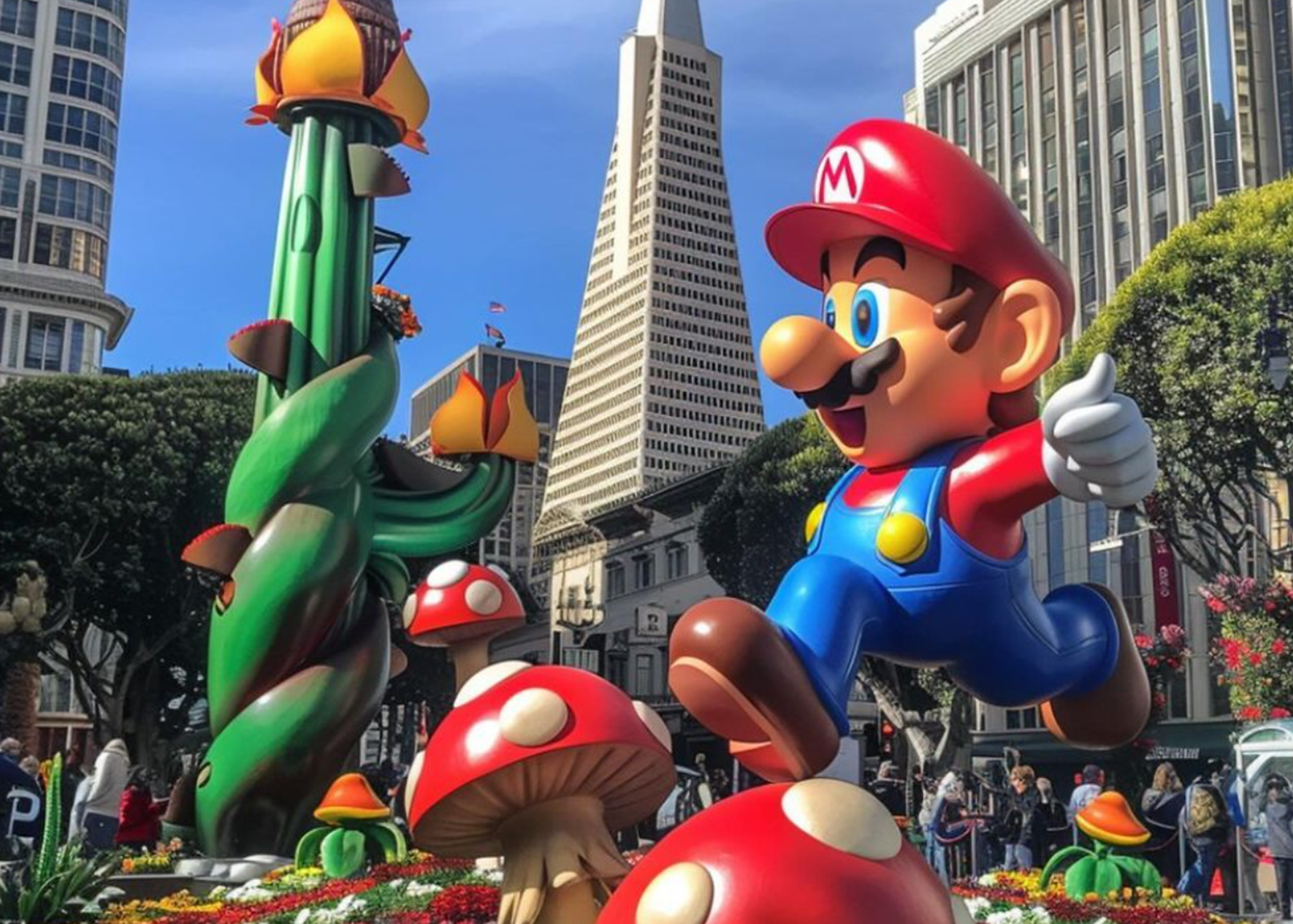 The image shows a large statue of a video game character dressed in red and blue, leaping with a thumbs up gesture, surrounded by oversized mushrooms and a green, twisting vine.