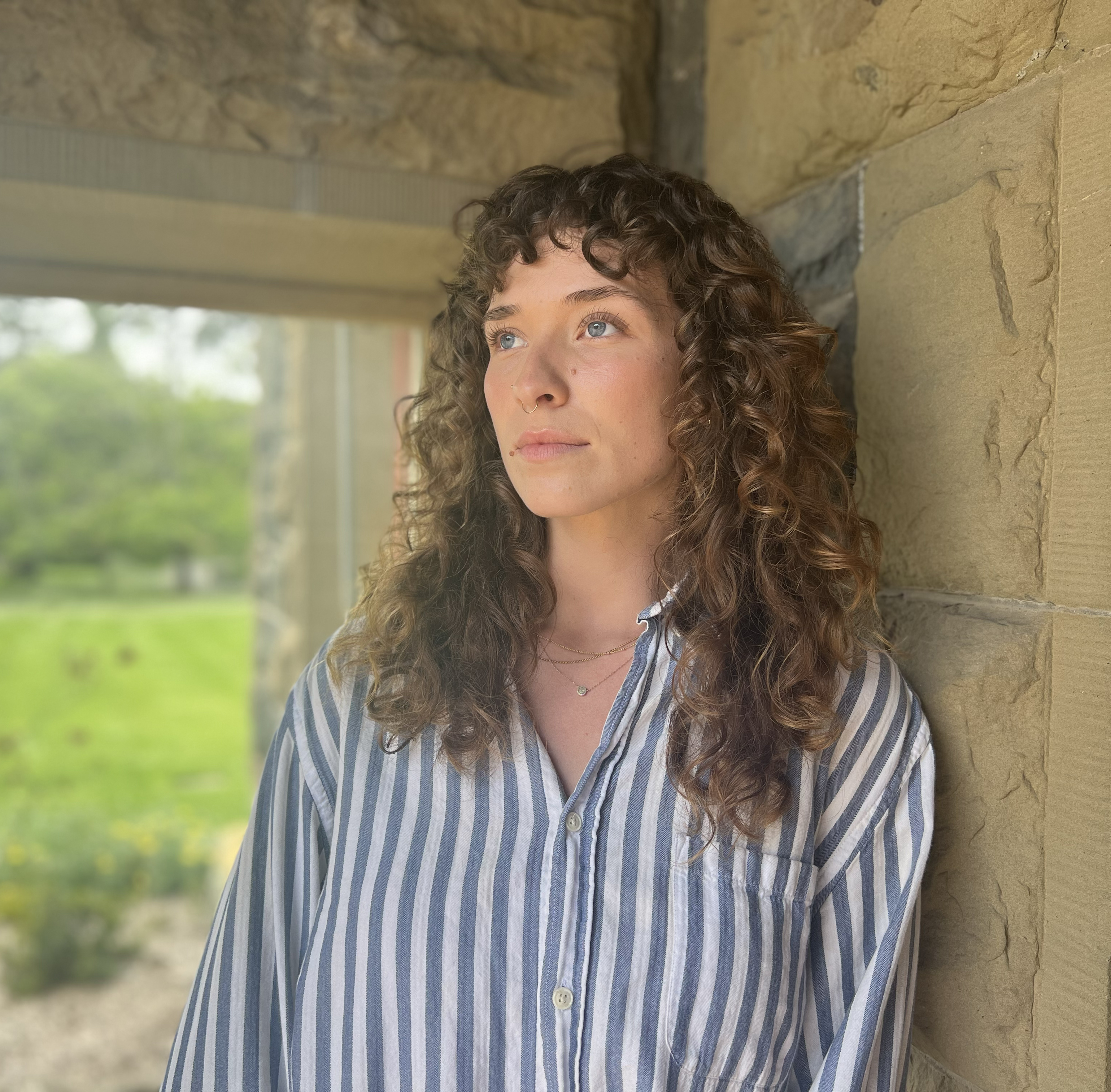 A woman with curly hair, wearing a striped shirt, stands under an archway, looking thoughtfully to the side, with greenery in the background.