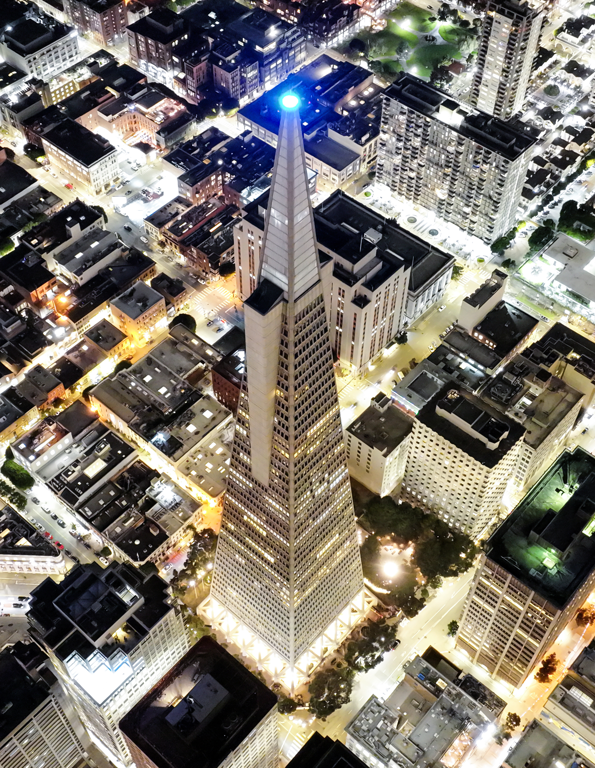 An aerial night view of a brightly lit, pyramid-topped skyscraper amid a grid of city streets.