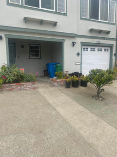 A house with a garage, a driveway, and a covered patio area with some potted plants. There are also blue and green recycling bins beside a small brick planter.