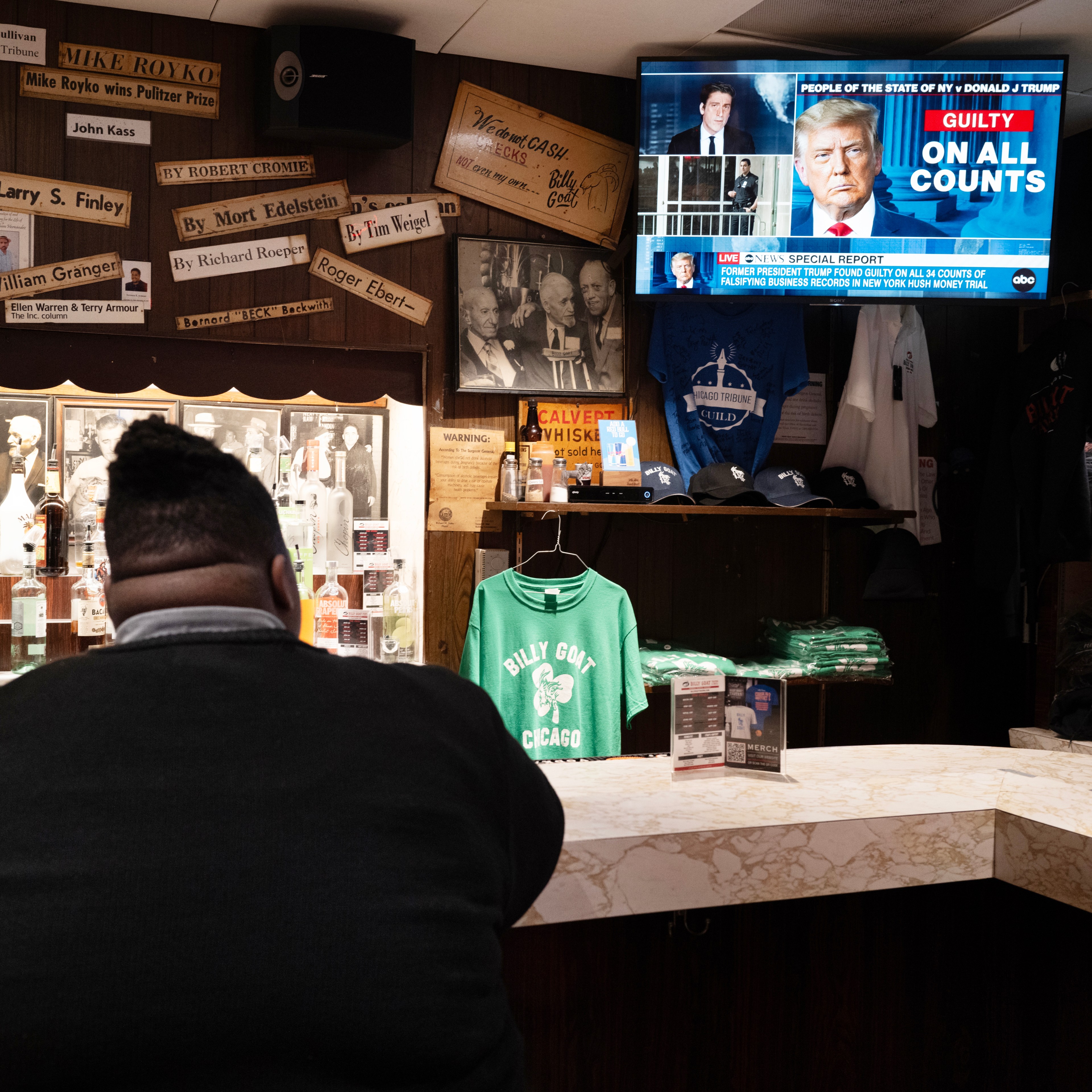 A person seated at a bar watches a TV showing news of Donald Trump found guilty on all counts. The bar has memorabilia and merchandise like hats and shirts.