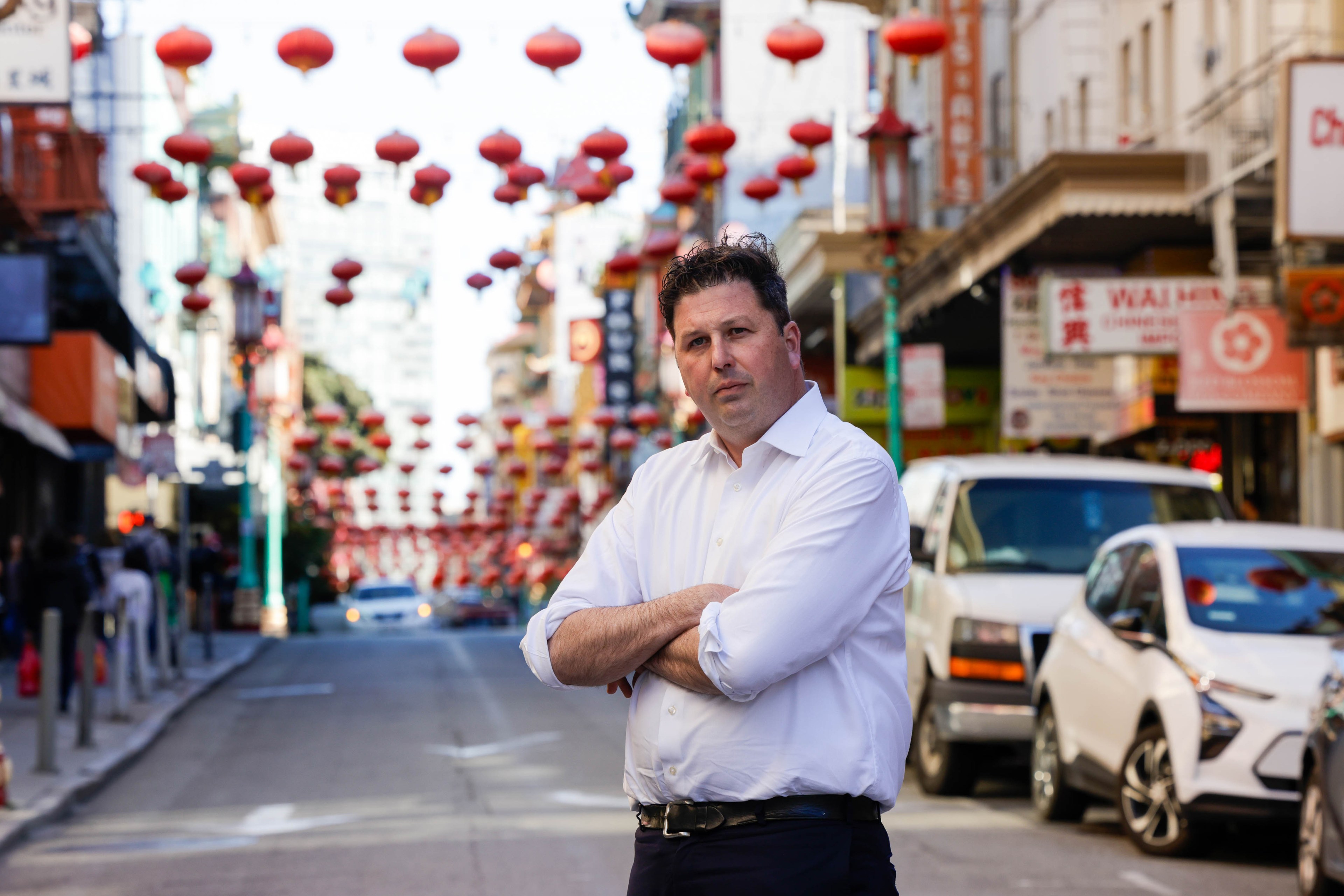A man stands with crossed arms on a street lined with red lanterns and signs, possibly in Chinatown.