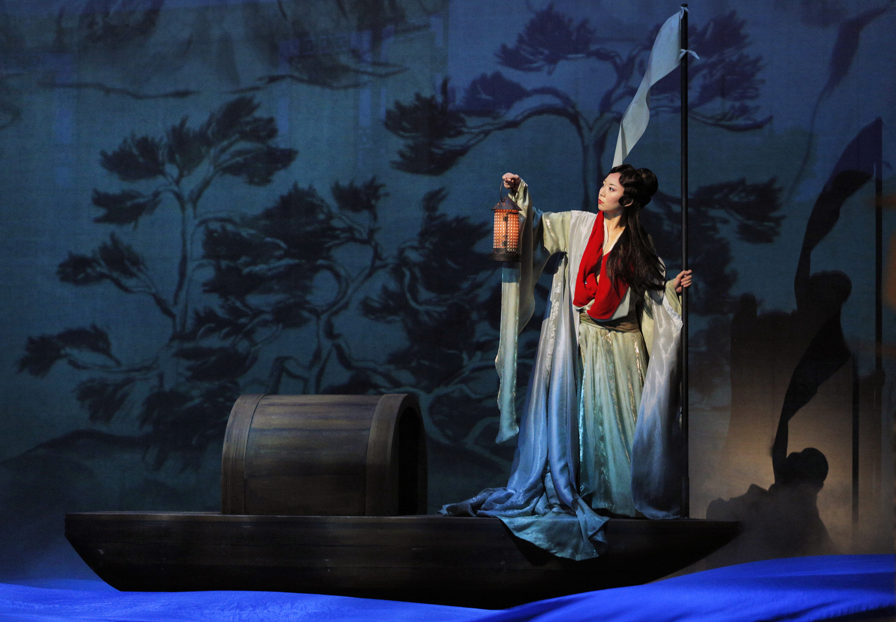 A woman in traditional East Asian attire holds a lantern while standing on a wooden boat with a blue-and-green painted backdrop of trees and mist.