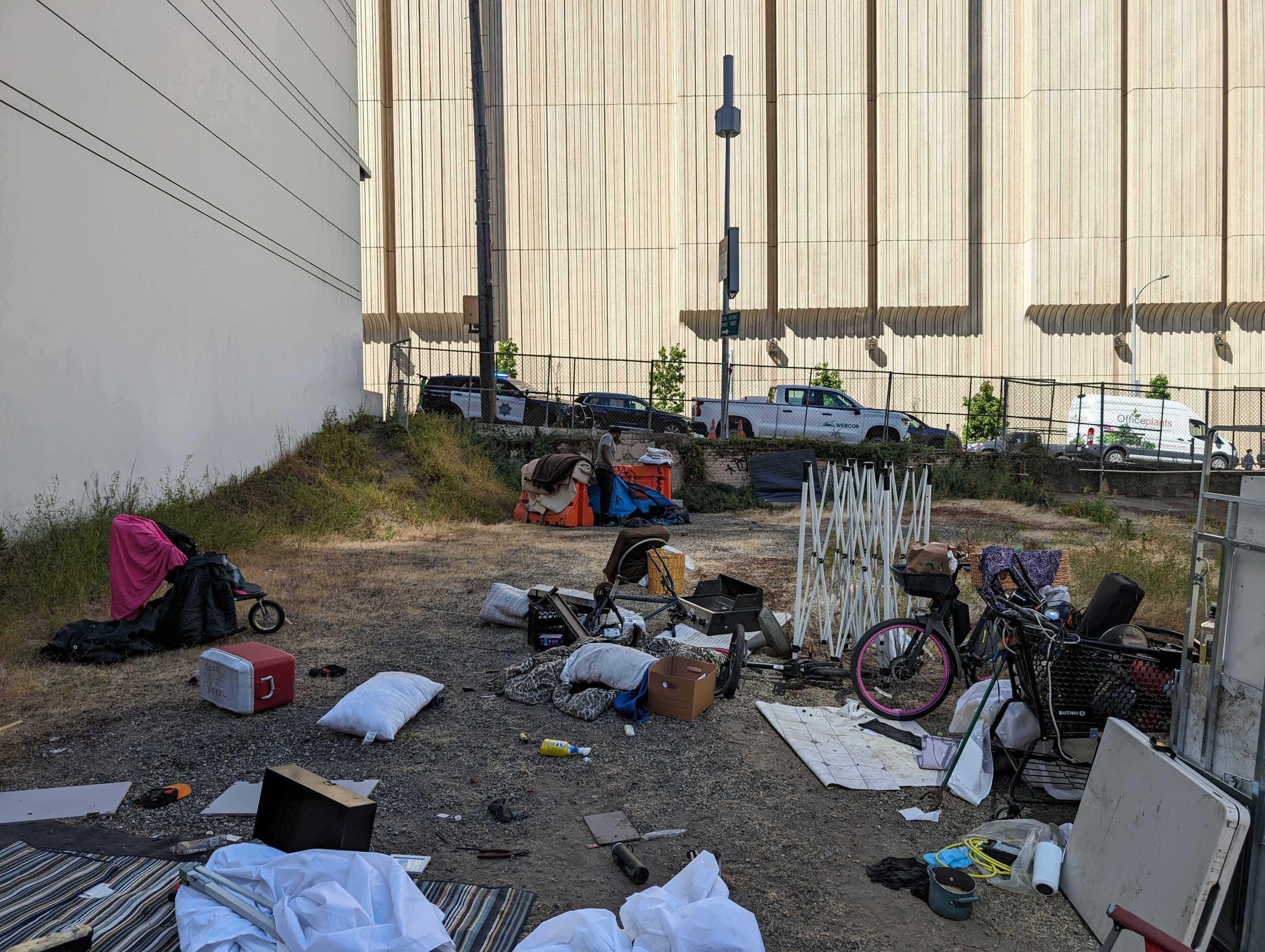 The image shows an outdoor area with scattered belongings including a tricycle, a cooler, pillows, a bike, and various items on the ground, suggesting a makeshift living space.