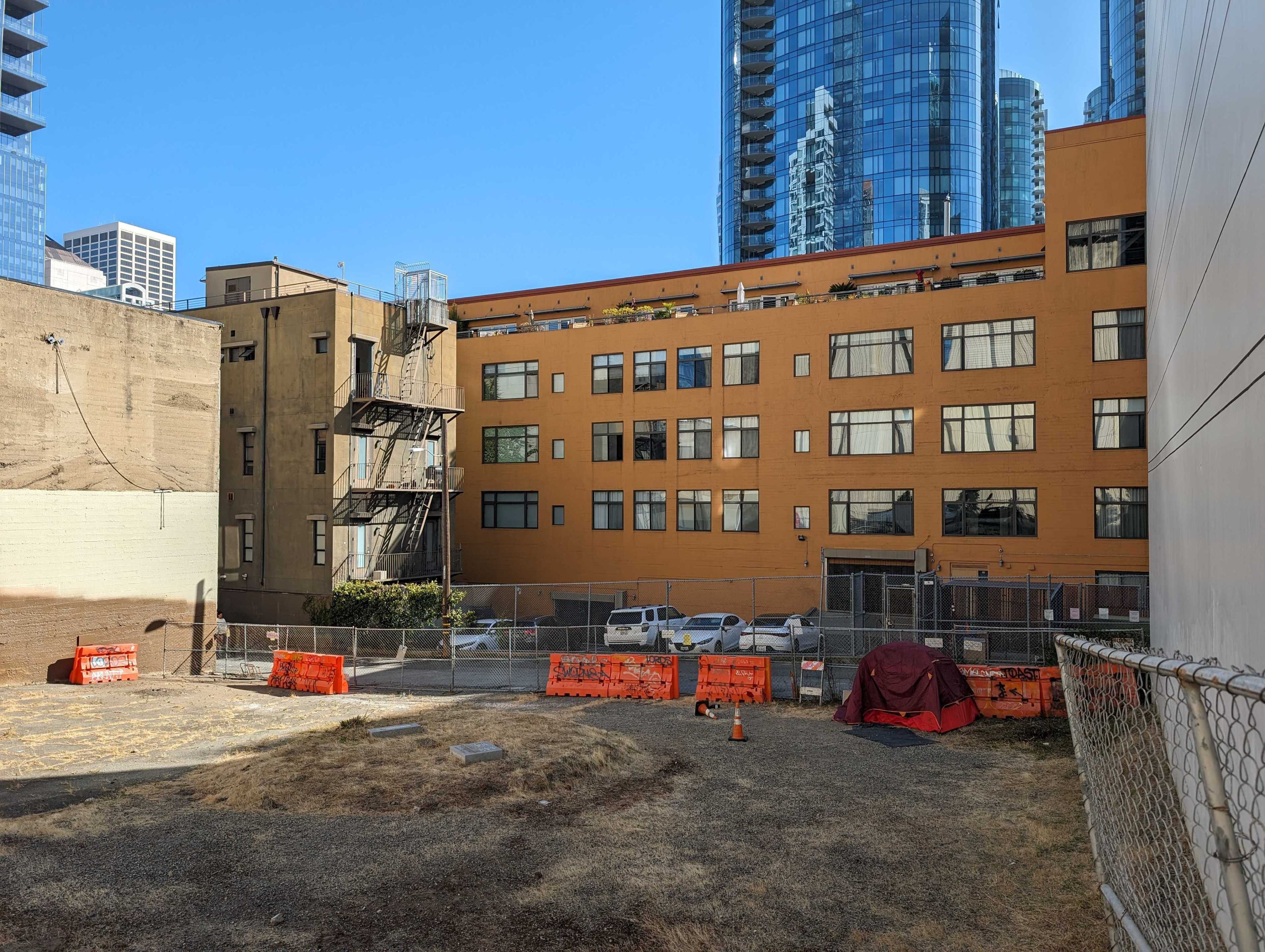 The image shows a vacant, fenced lot surrounded by tall buildings, including a beige and orange building. Safety barriers and a makeshift tent are visible.
