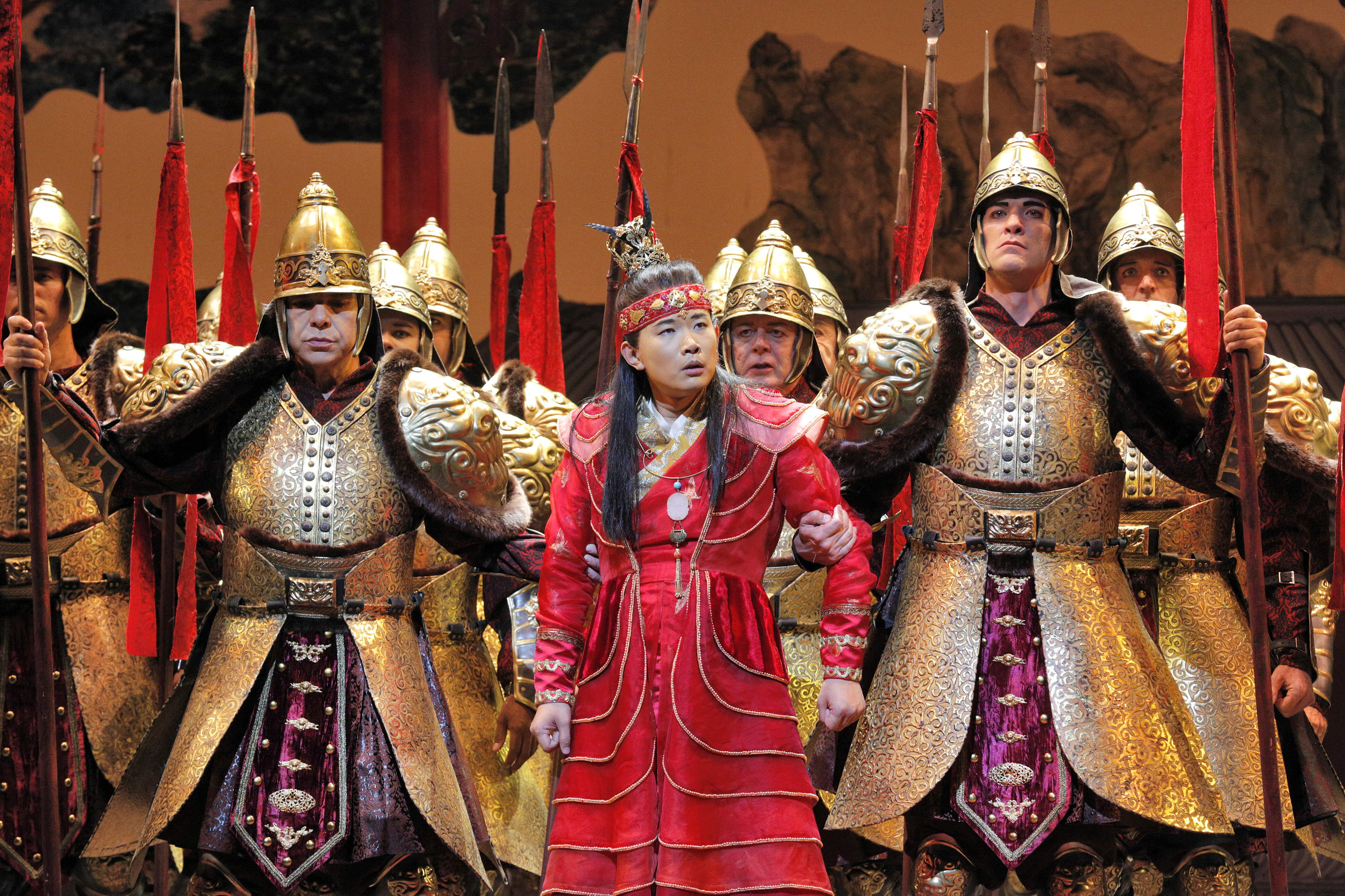 A person in red historical attire and a crown stands, looking surprised, surrounded by armed soldiers in ornate gold armor holding spears with red tassels.