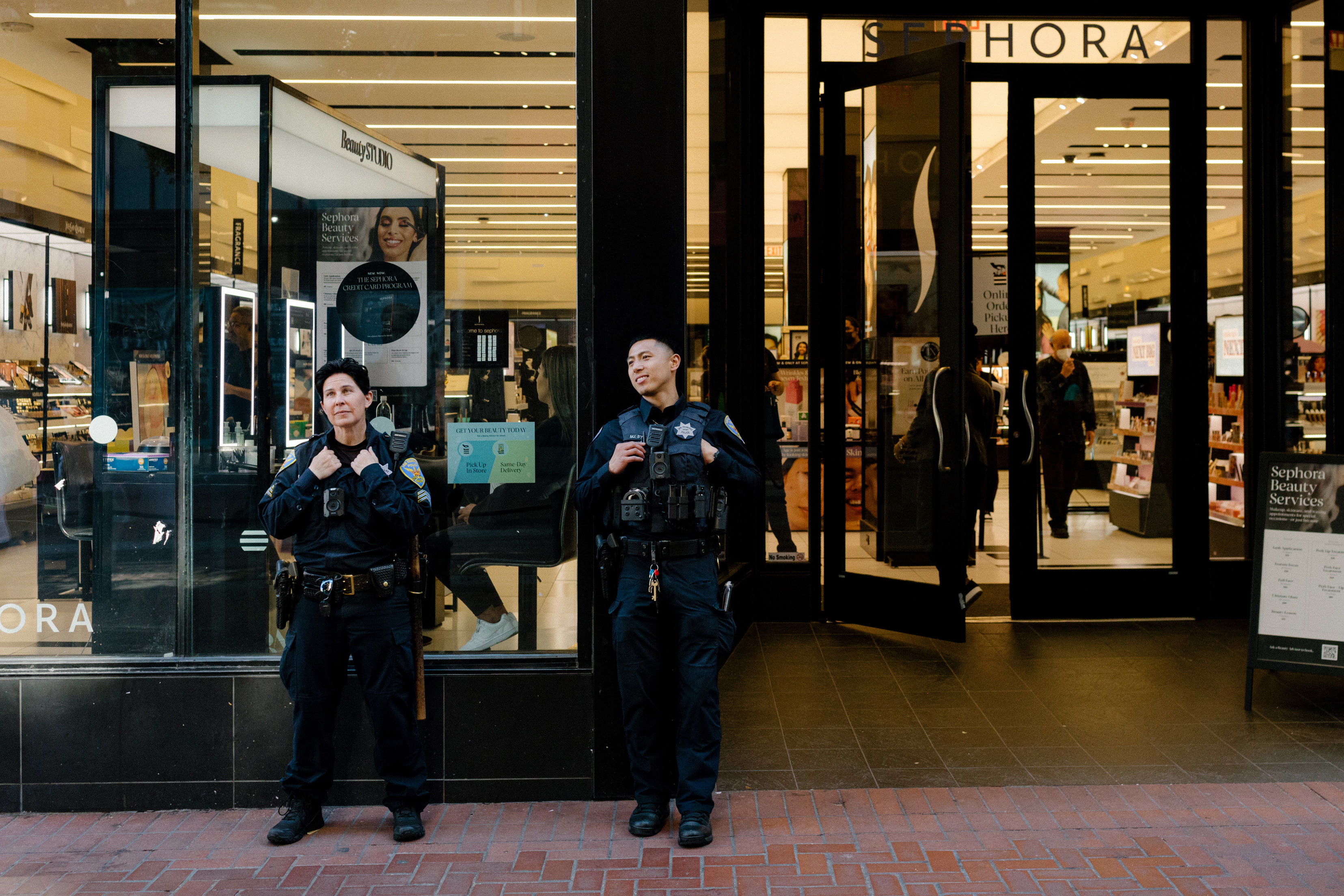 Two police officers, in uniform with vests, stand outside a Sephora store; one officer looks ahead, while the other looks upward. The store entrance is open.