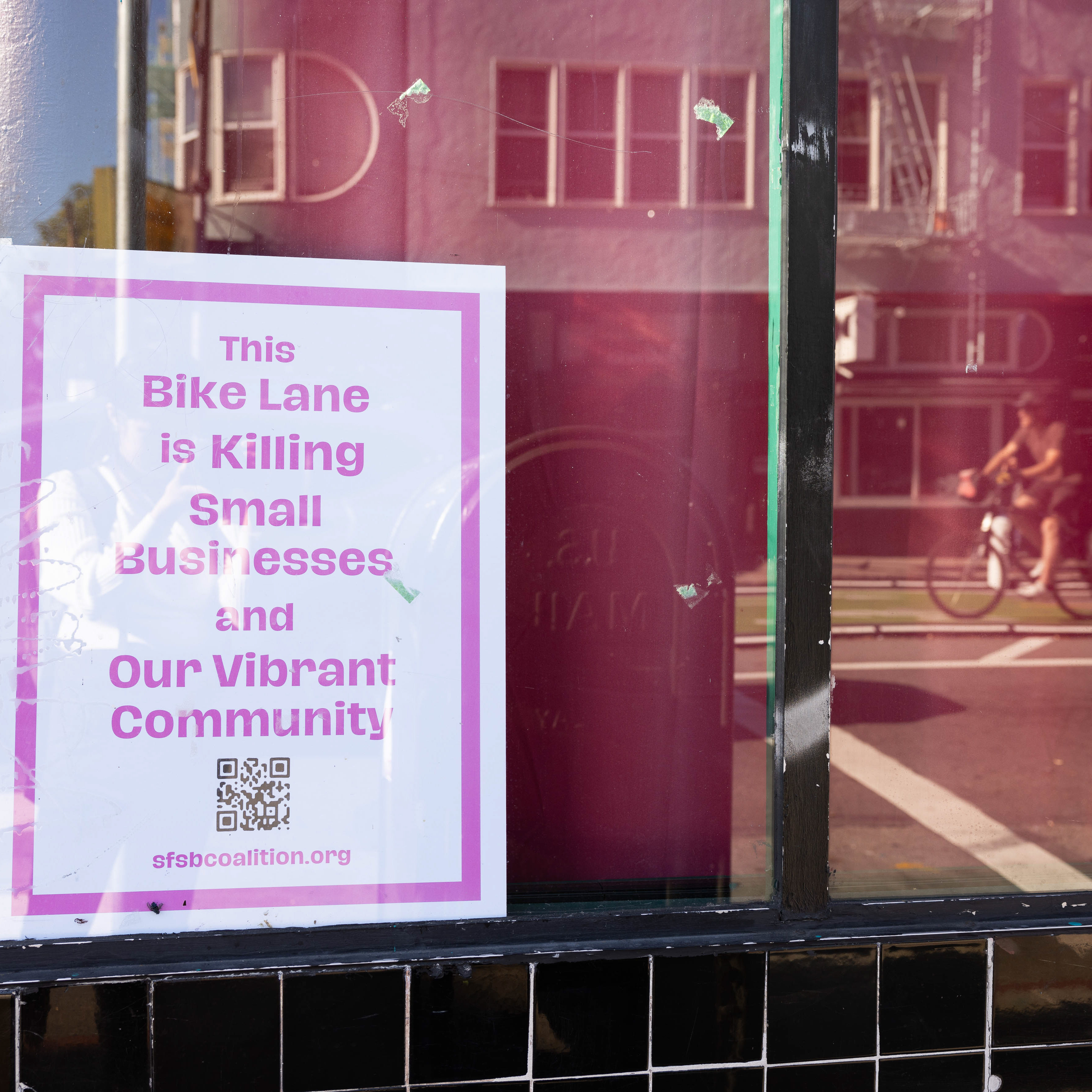 A sign in a shop window reads, &quot;This Bike Lane is Killing Small Businesses and Our Vibrant Community,&quot; with a QR code and an organization URL at the bottom.