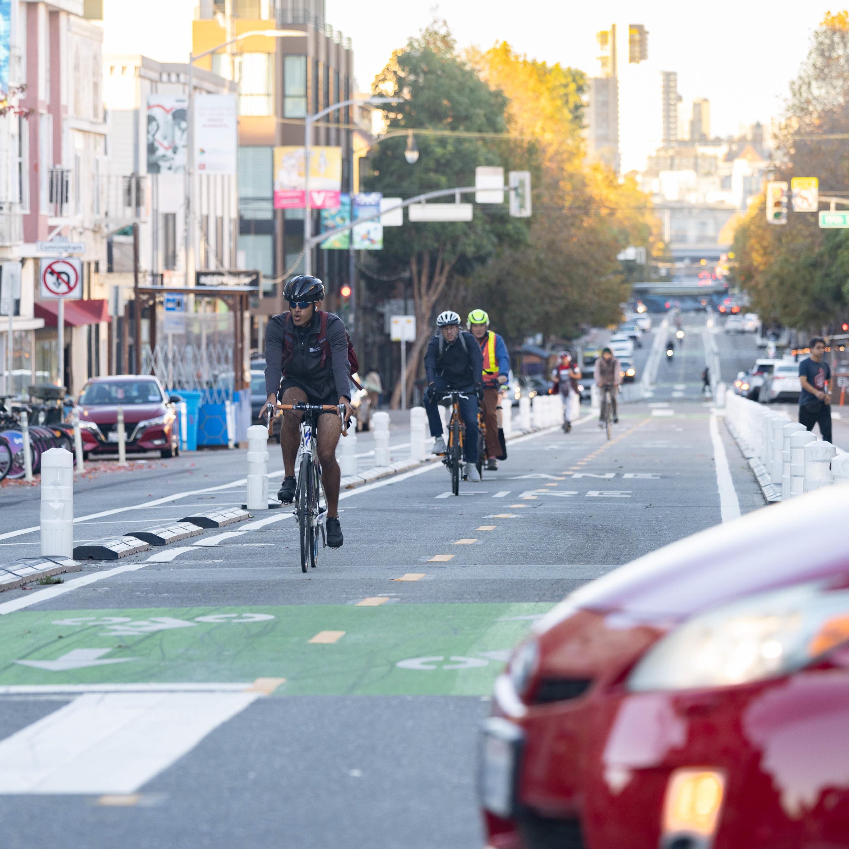 This image shows a city street with several cyclists riding in a dedicated bike lane, separated from cars by plastic bollards. In the background, there are buildings and trees.