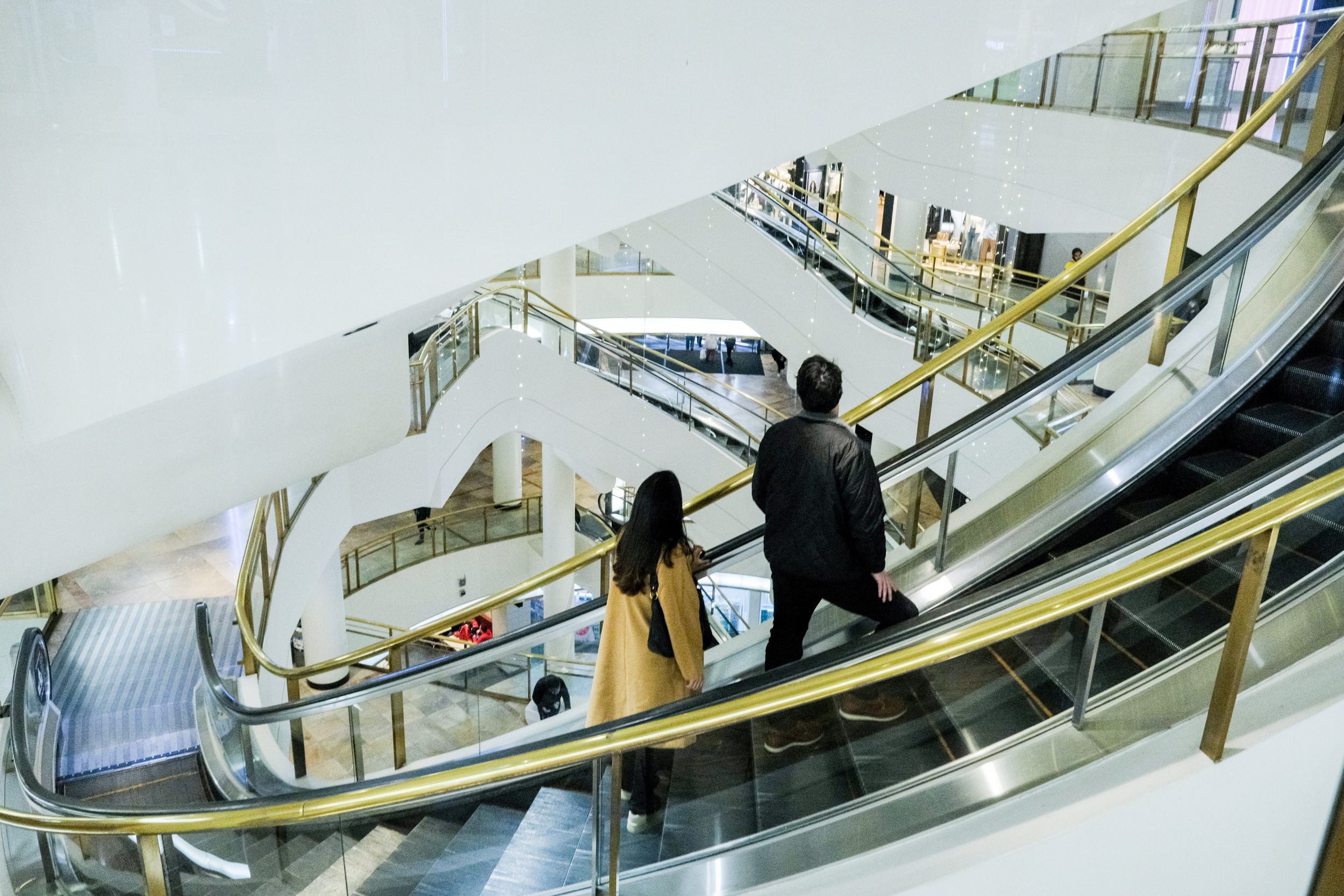 Two people, one in a black jacket and the other in a yellow coat, are on an ascending escalator in a modern multi-level shopping mall with white interiors and glass railings.