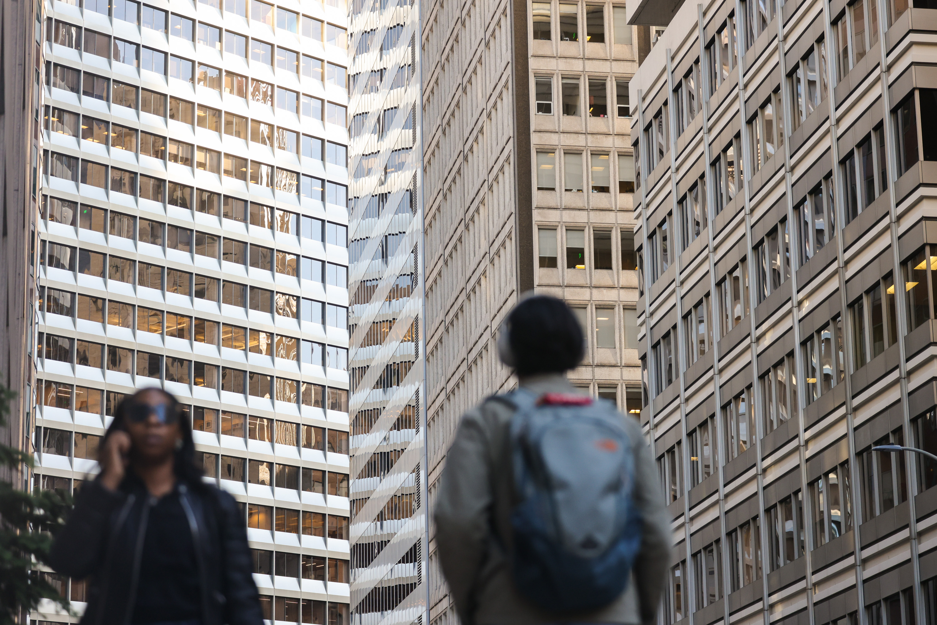 Two people walk in front of tall office buildings, one person wearing sunglasses and using a phone, the other carrying a backpack.