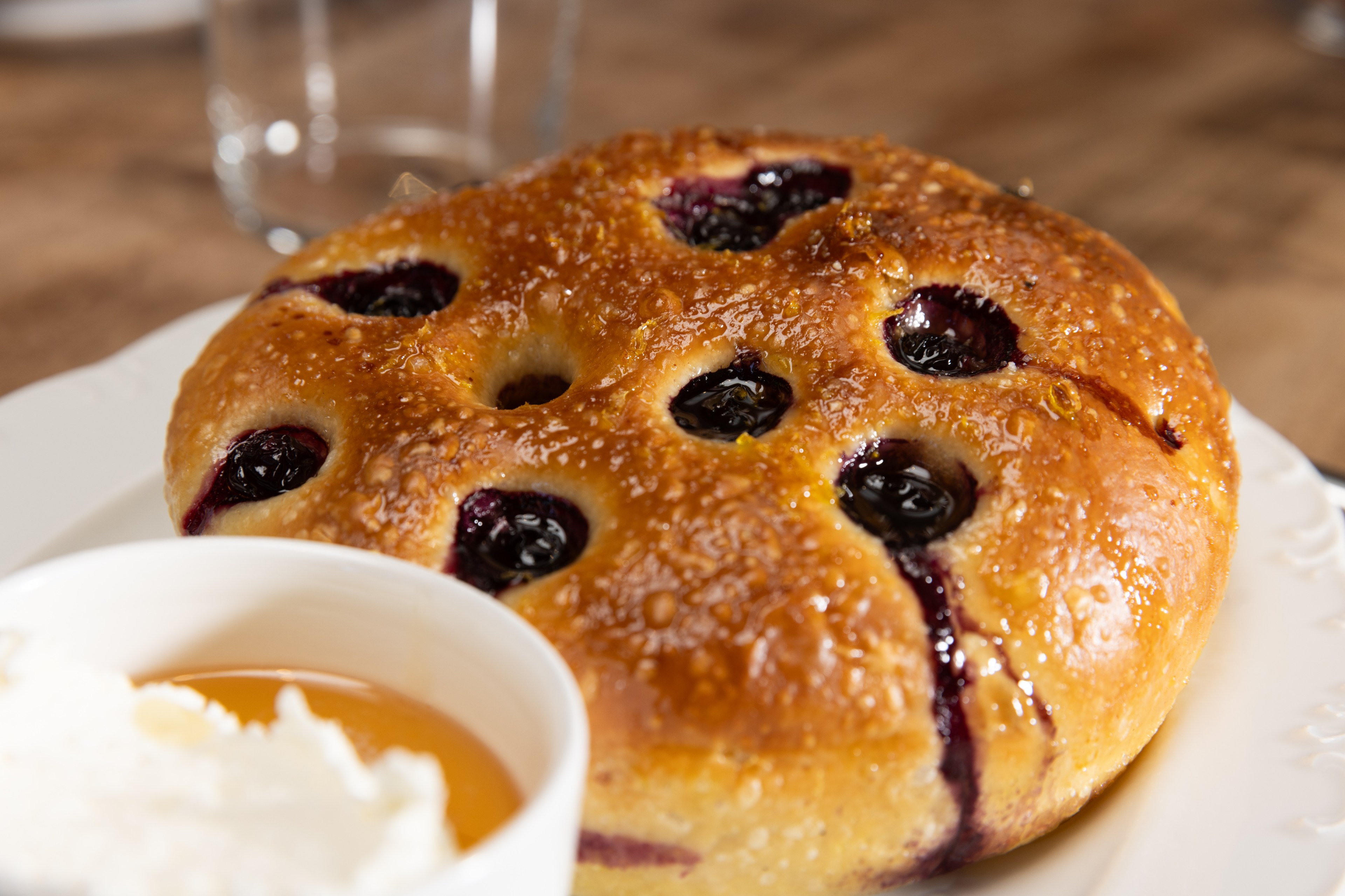 A golden-brown pastry dotted with juicy blueberries sits on a white plate, accompanied by a bowl of whipped cream and syrup in the foreground.