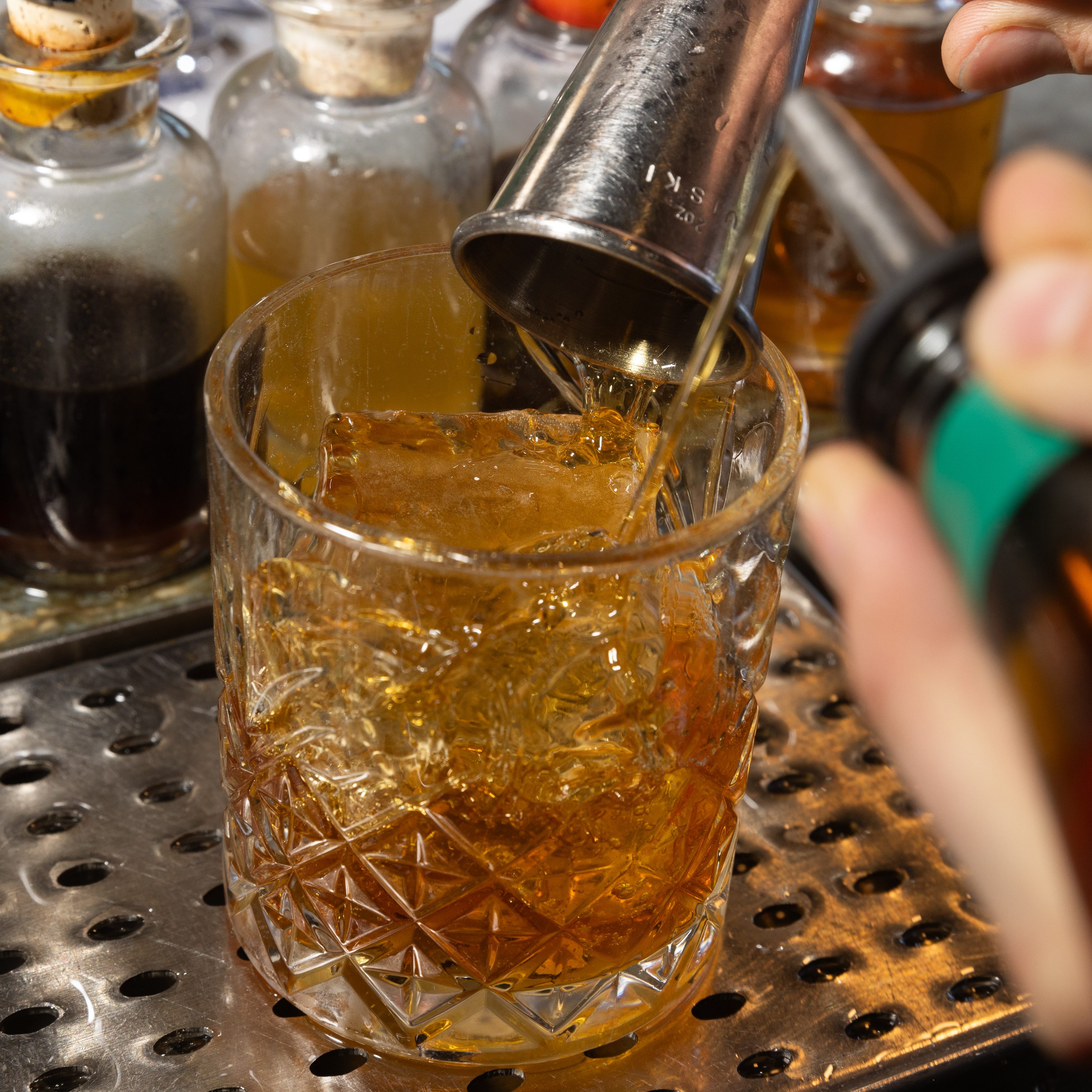 A hand is pouring a golden liquid from a jigger into a glass with ice on a metal drip tray. Several small glass bottles with cork stoppers are visible in the background.