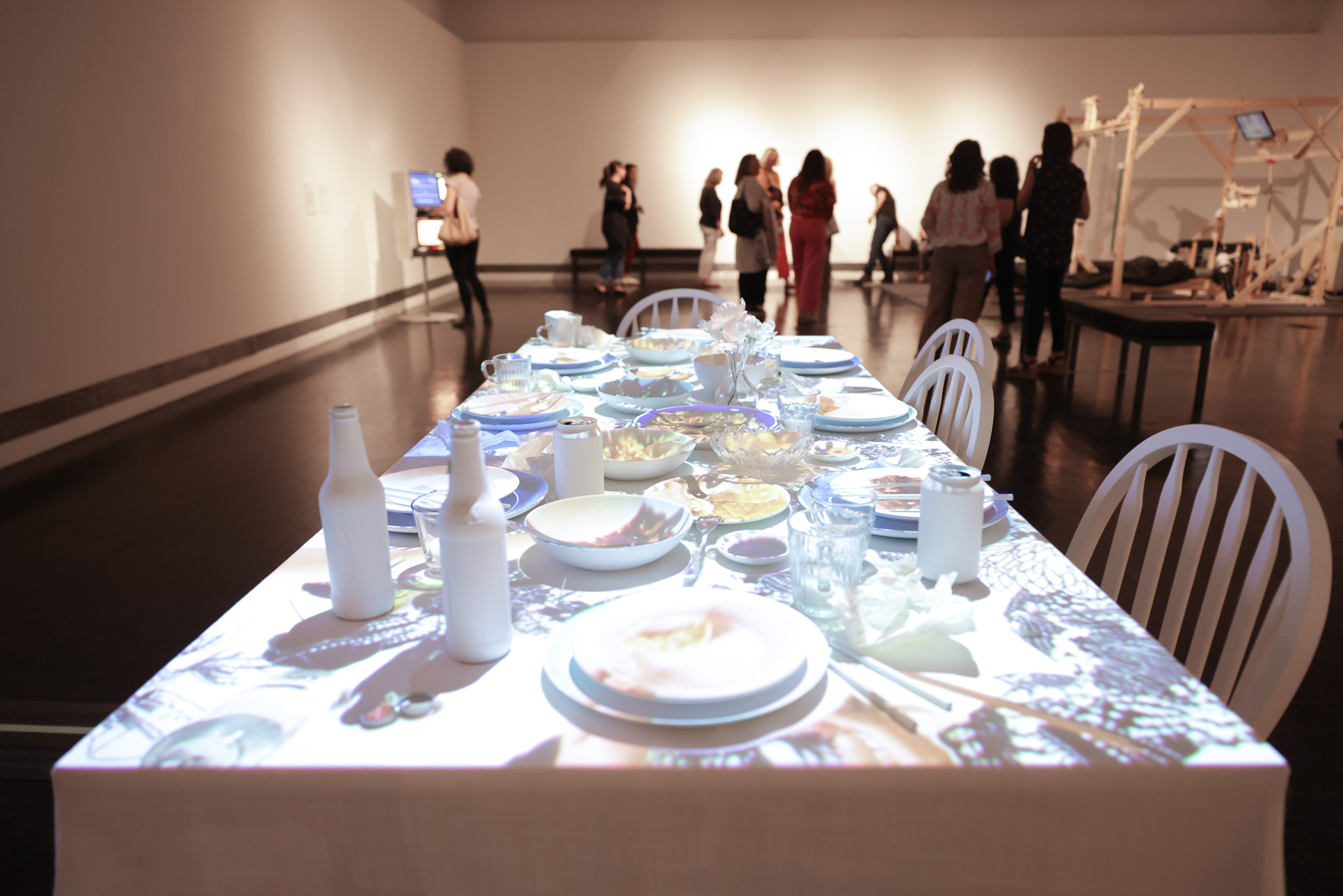 The image shows a well-set dining table with white plates, bottles, and cups, while a group of people stand and walk in the background in a spacious room.
