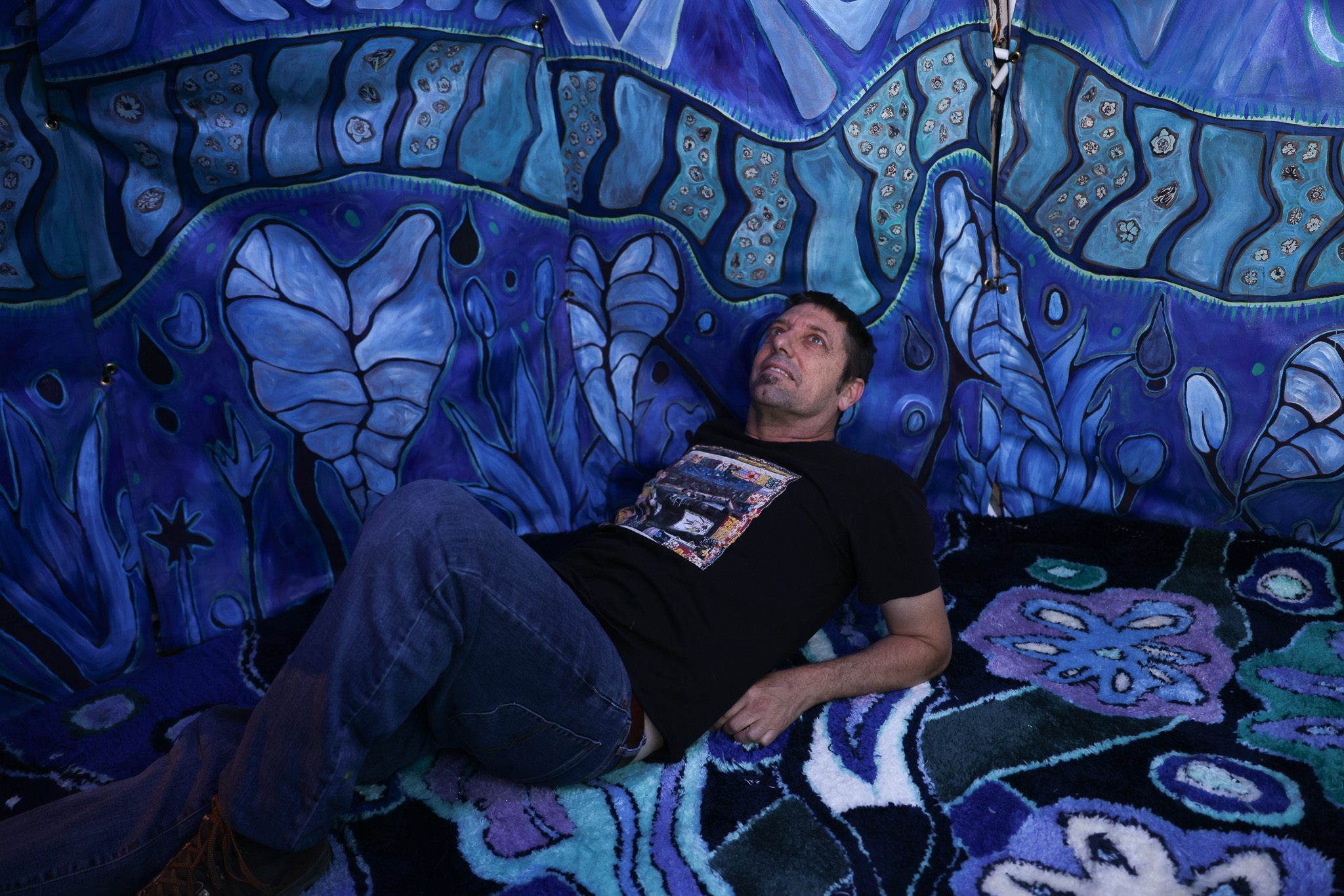 A man in a black T-shirt and jeans lies on a colorful, patterned carpet with his head resting against a vibrant blue wall adorned with abstract designs.