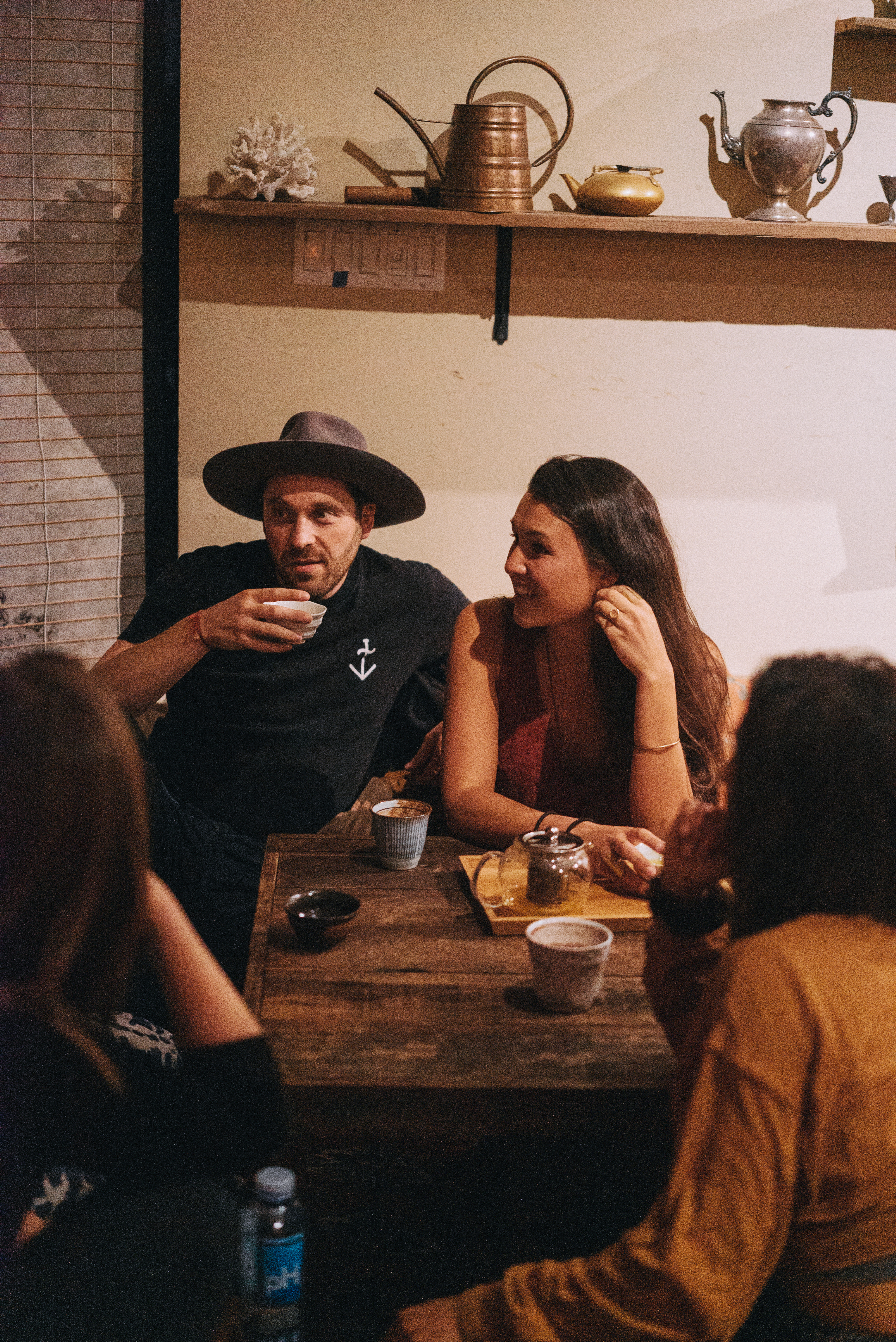Four people sit around a wooden table with tea cups and a teapot, engaged in conversation. One man wears a hat and drinks from a cup, while a woman smiles beside him.