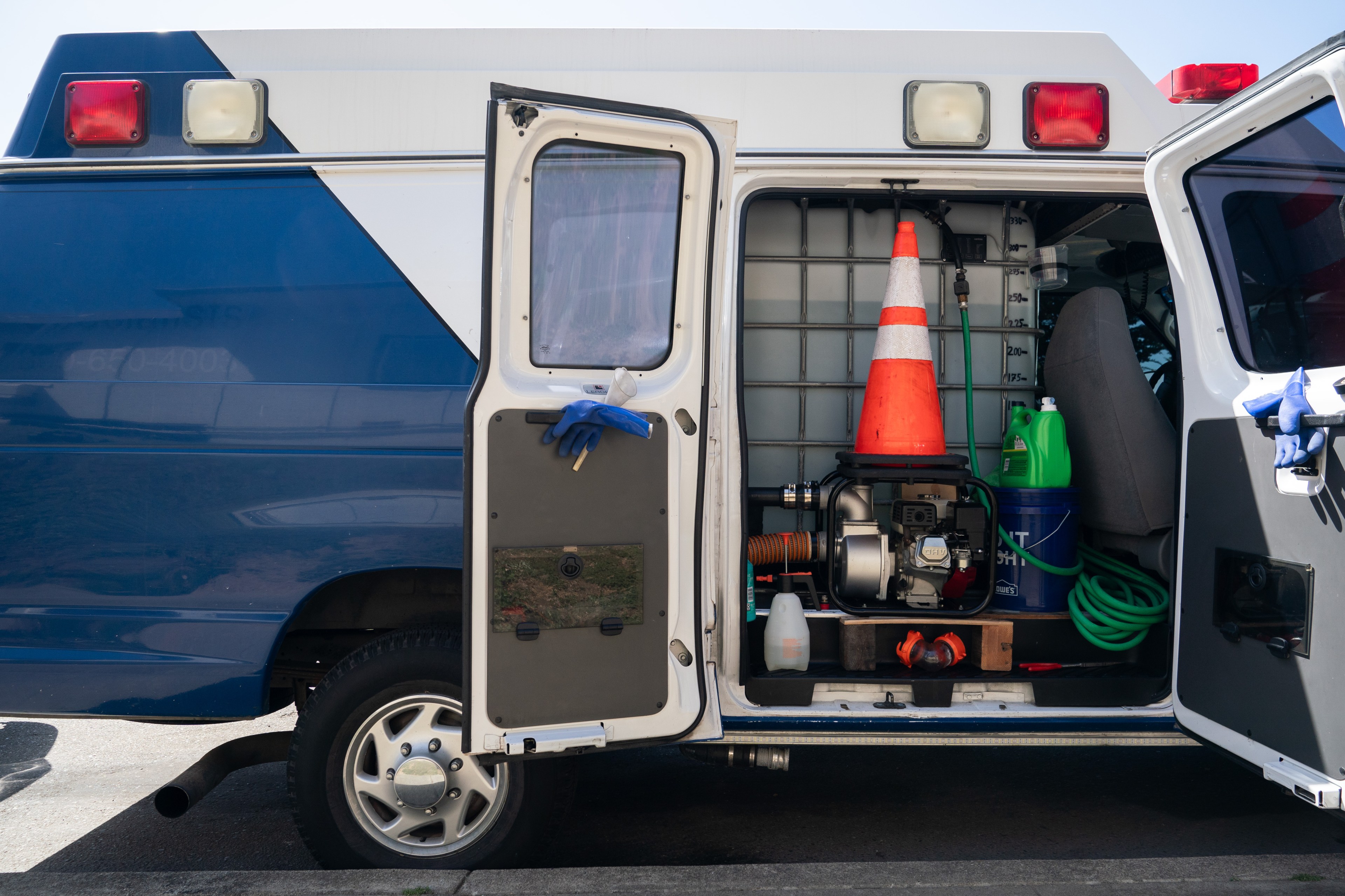 The back doors of a blue and white emergency vehicle are open, revealing equipment such as a traffic cone, green hose, motor, and cleaning supplies.