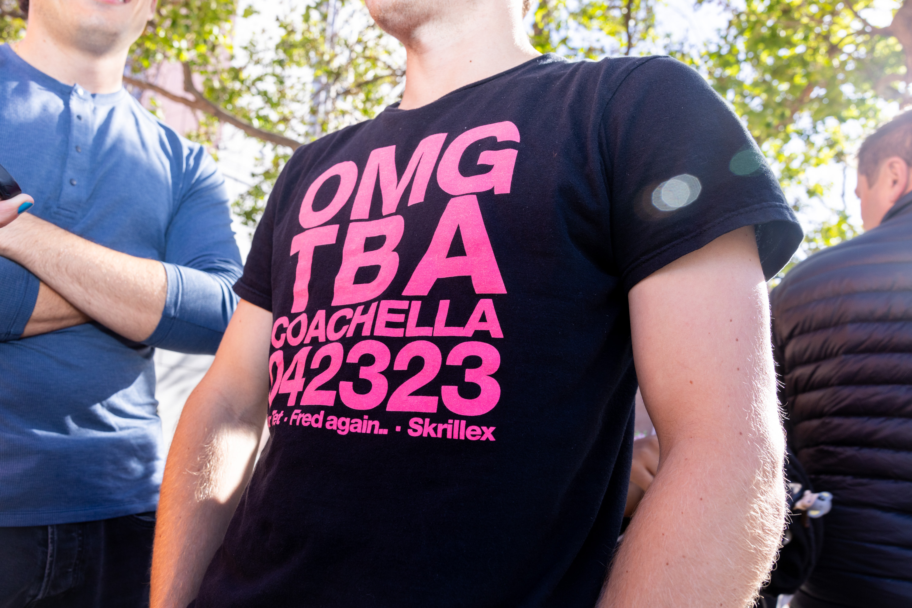 A person wears a black T-shirt with large, bright pink text promoting Coachella. The text reads, &quot;OMG TBA COACHELLA 042323,&quot; featuring &quot;Fred again..&quot; and &quot;Skrillex.&quot;