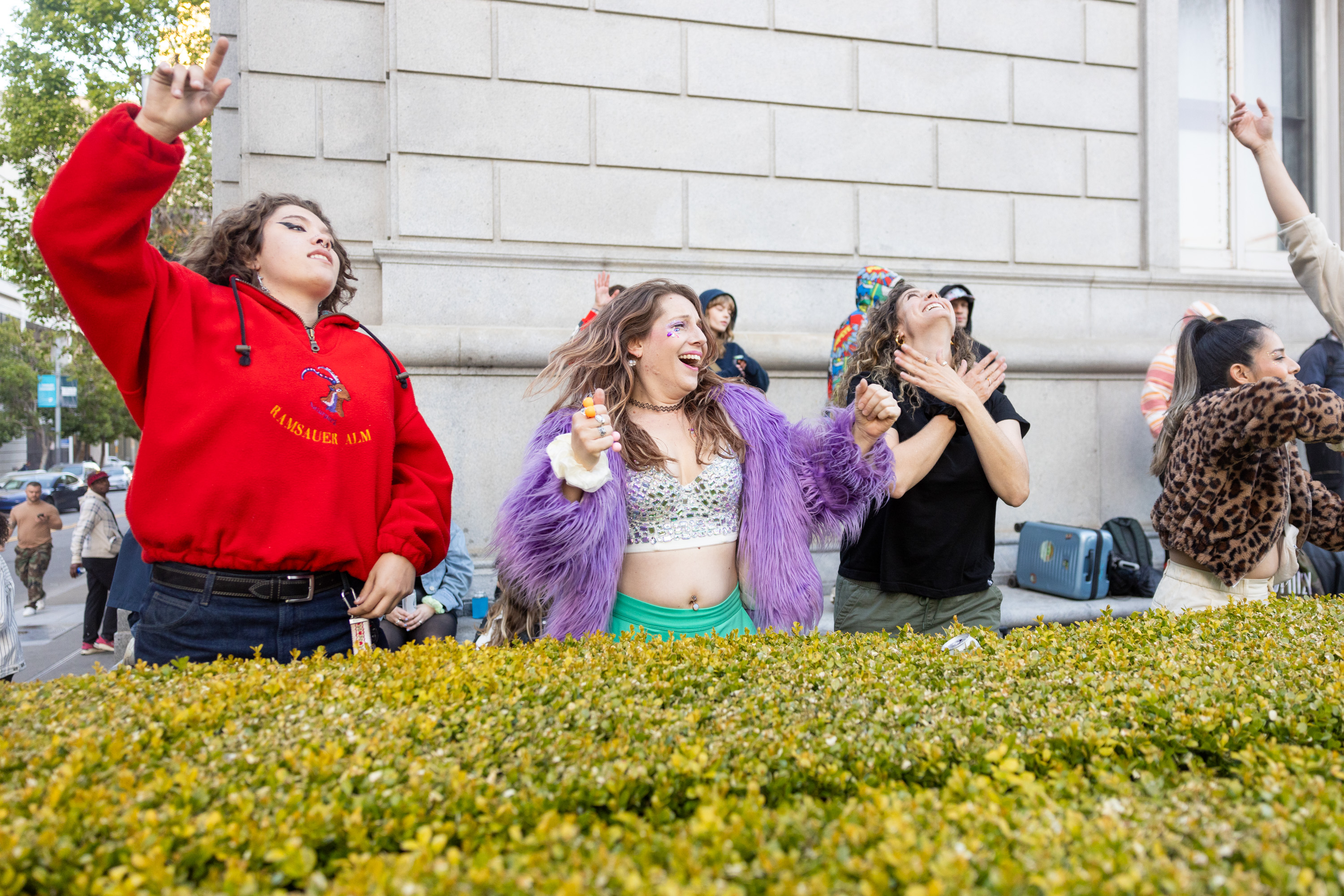 A group of joyous people dance energetically outside a large concrete building, some in colorful, furry coats, in front of green shrubbery.
