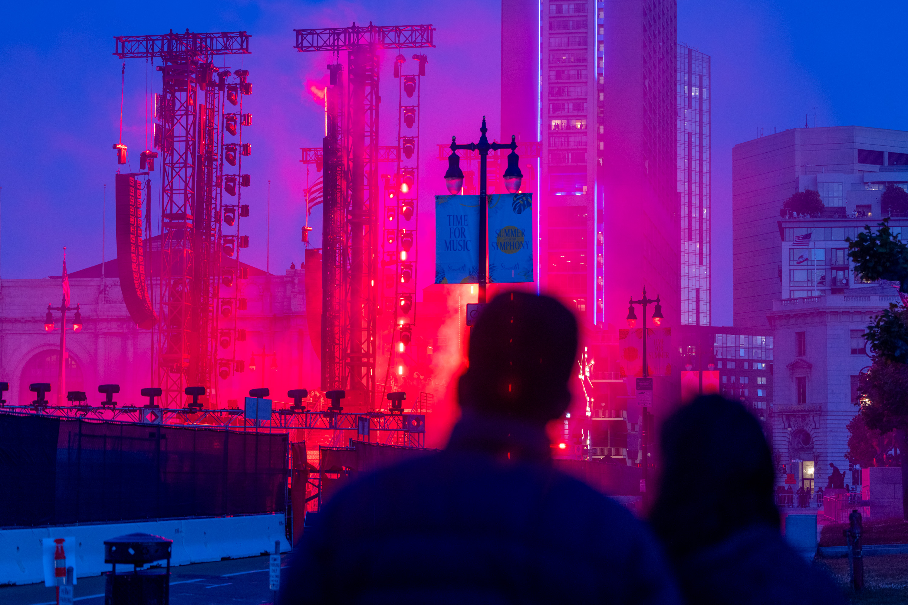 Two figures face a vibrant urban scene with bright red lights, stage equipment, and tall buildings at dusk. Banners read &quot;TIME FOR MUSIC&quot; and &quot;SUMMER SYMPHONY.&quot;