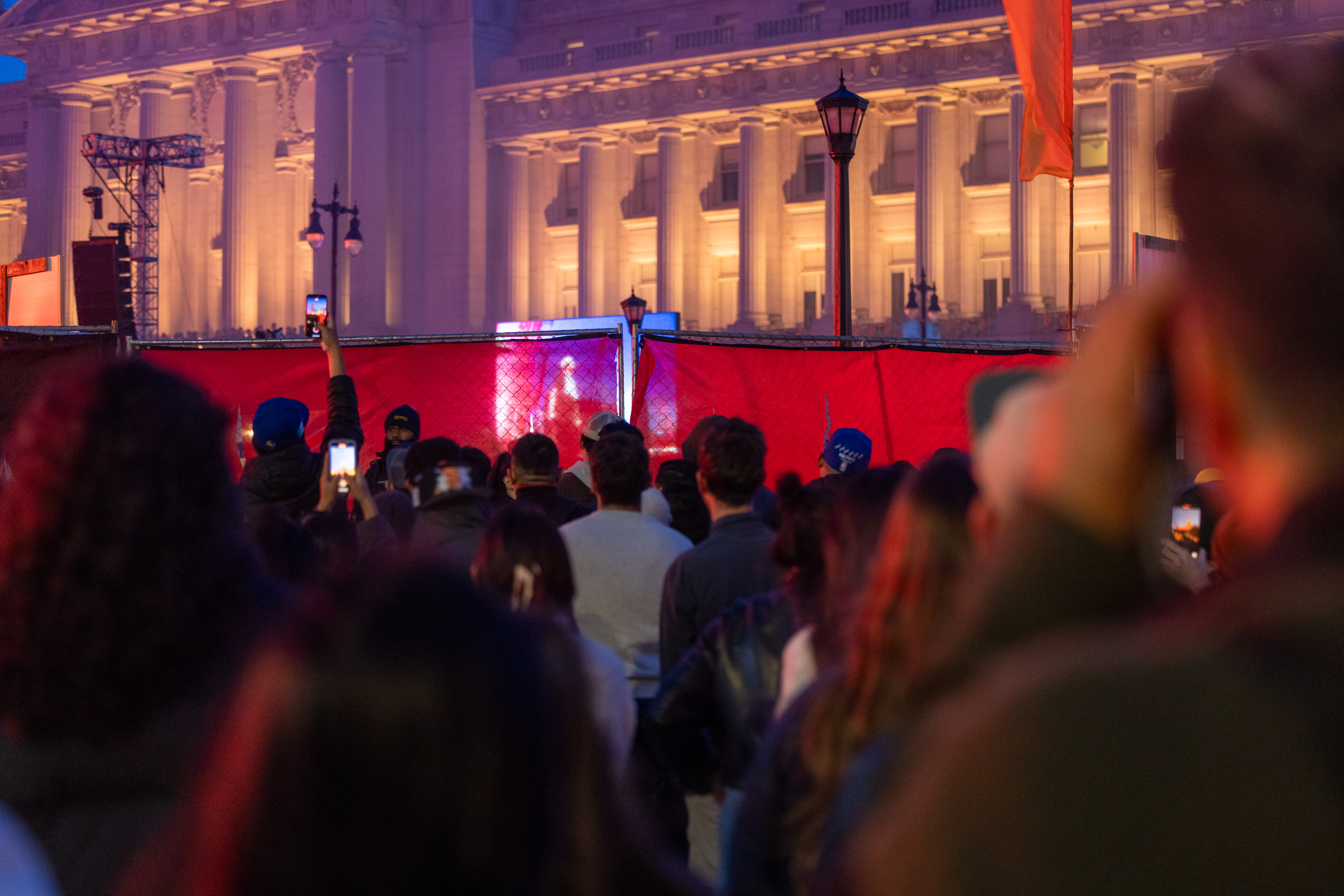 A crowd gathers in front of a lit, grand building at night. They are looking through a red barrier, holding up phones to capture photos or videos of something unseen.