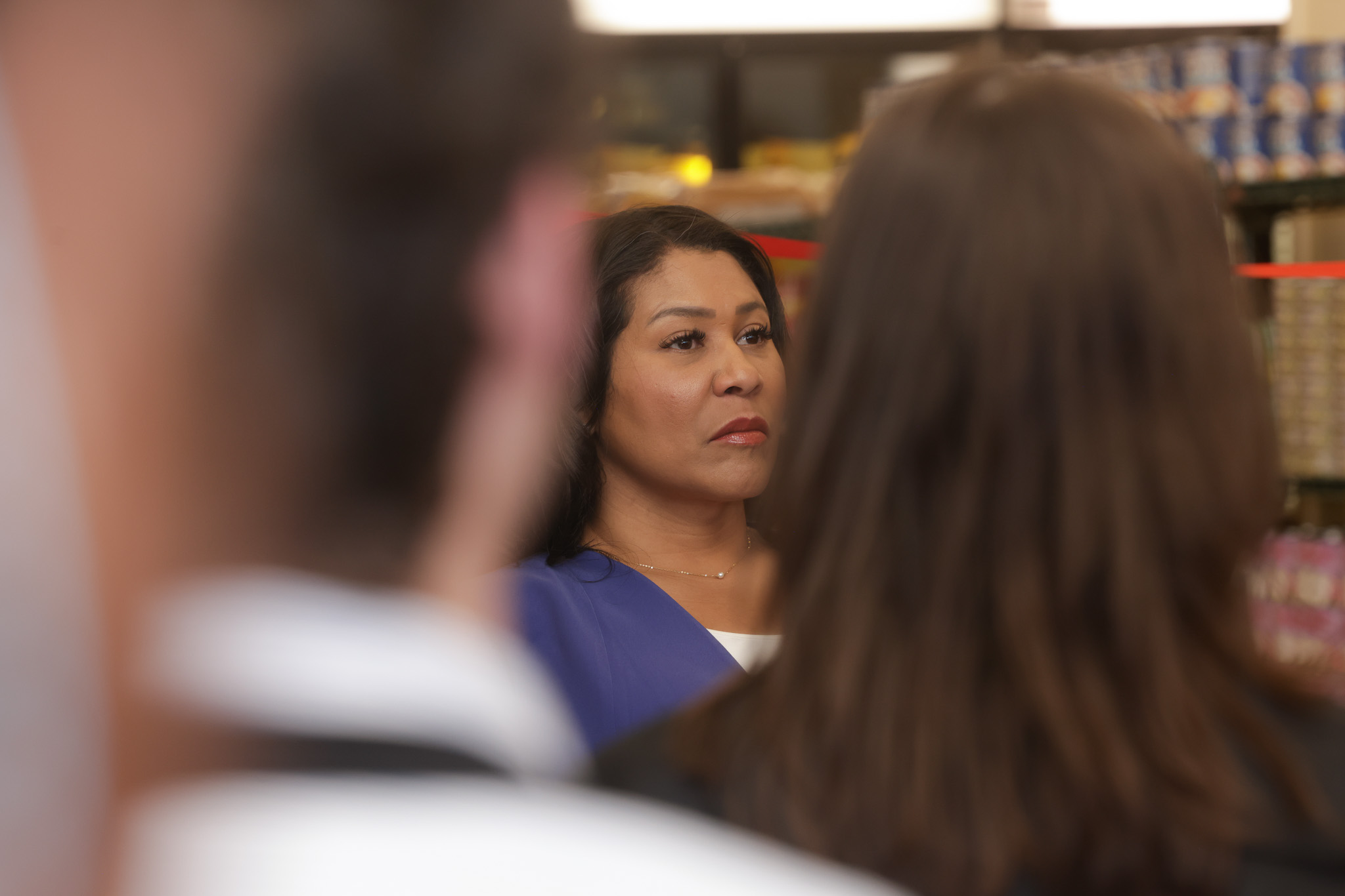 A woman with a serious expression stands in focus, surrounded by blurred figures in the foreground, against a backdrop of store shelves.