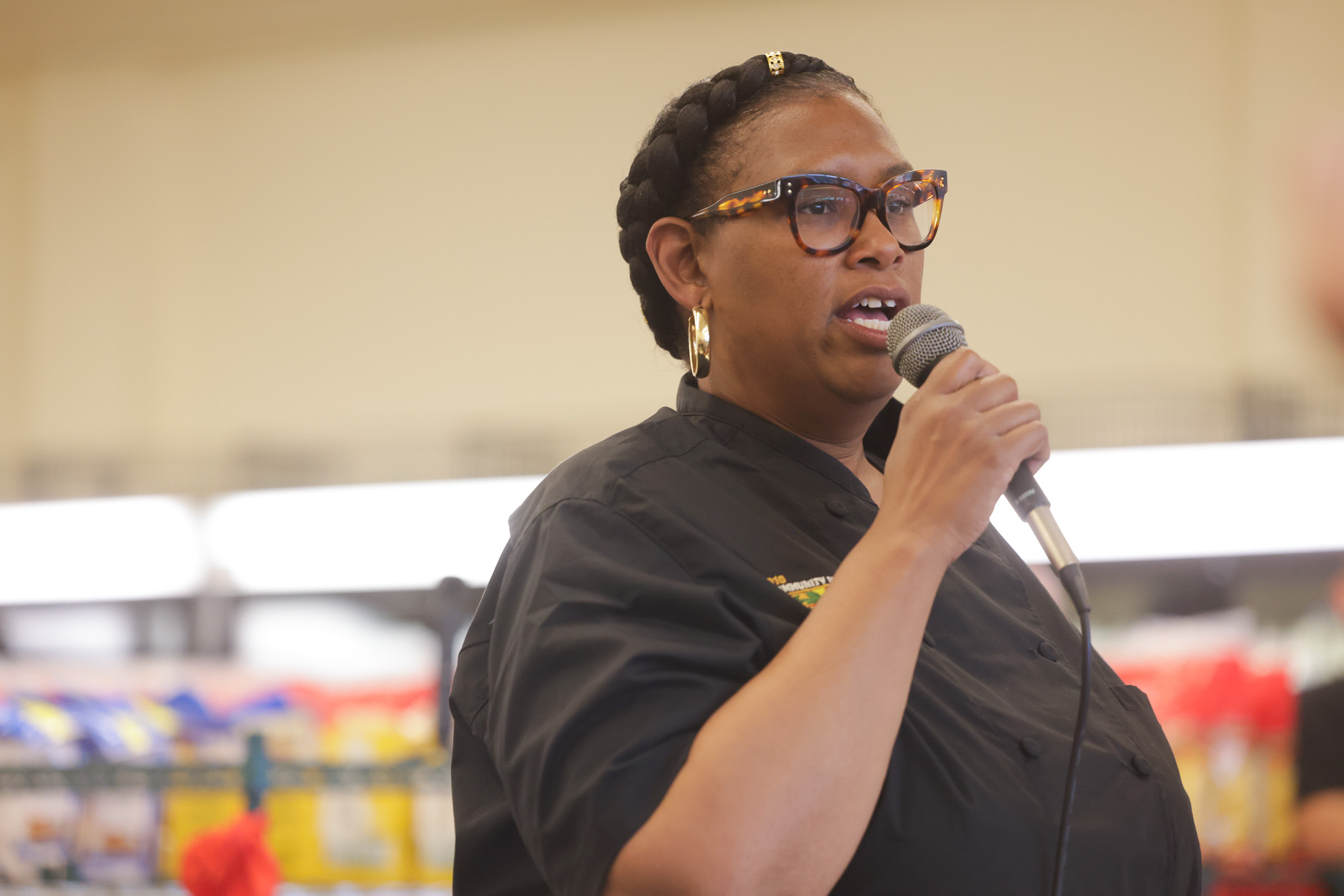 A person with short, braided hair and large, round glasses is speaking into a microphone. They are wearing a dark button-up shirt, possibly a uniform, in a brightly lit indoor space.