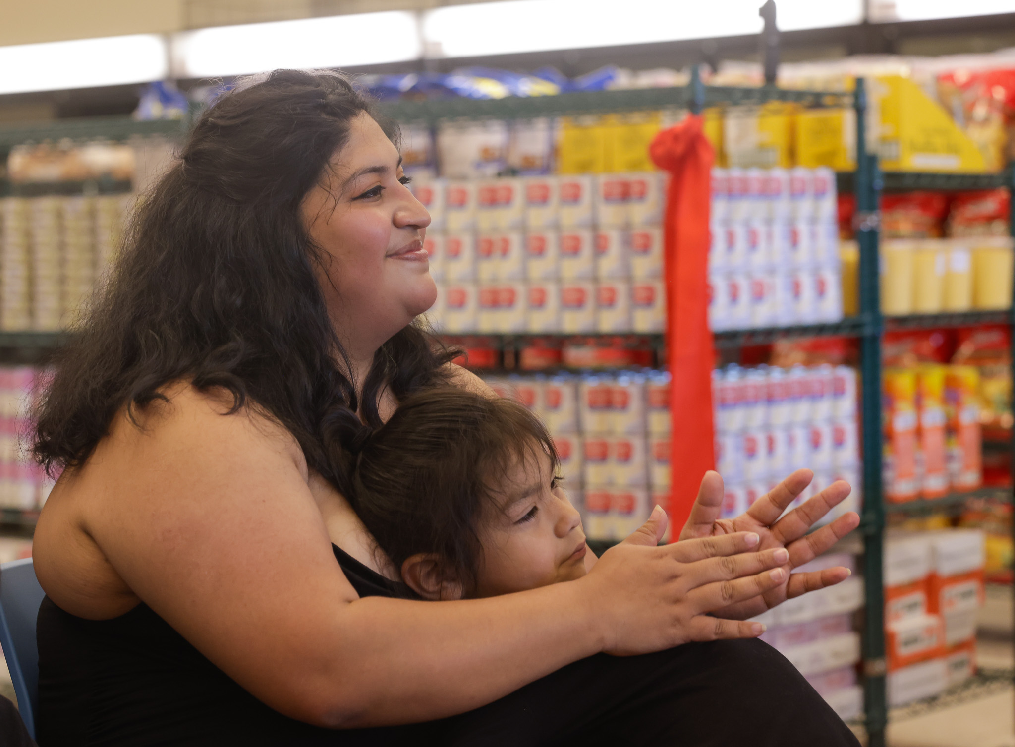 A woman and child are sitting in a store, clapping and smiling. The background shows shelves stocked with various products, including boxes and cans.