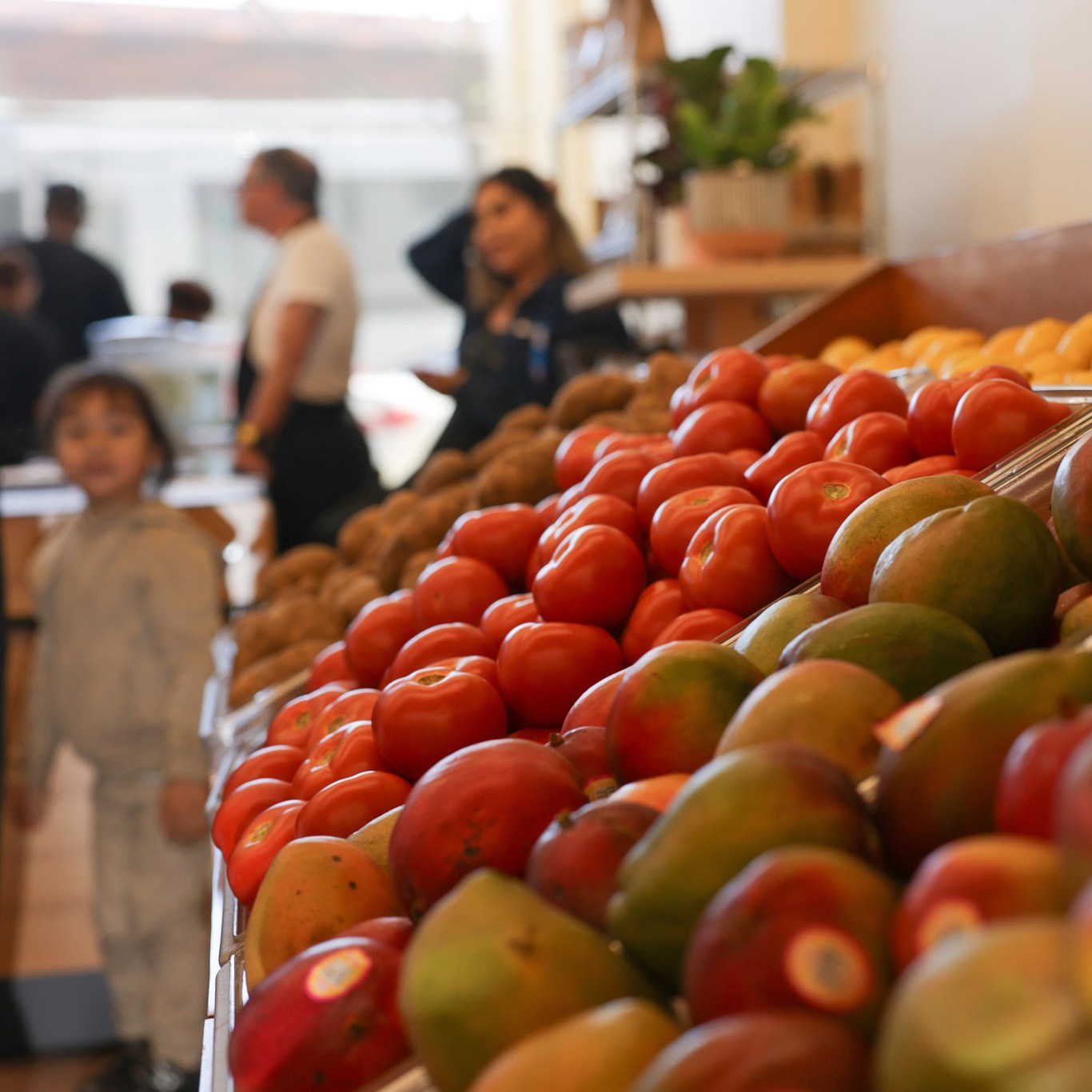 In a grocery store, colorful produce like tomatoes and mangoes are neatly displayed on a shelf, with people and a small child visible in the blurry background.