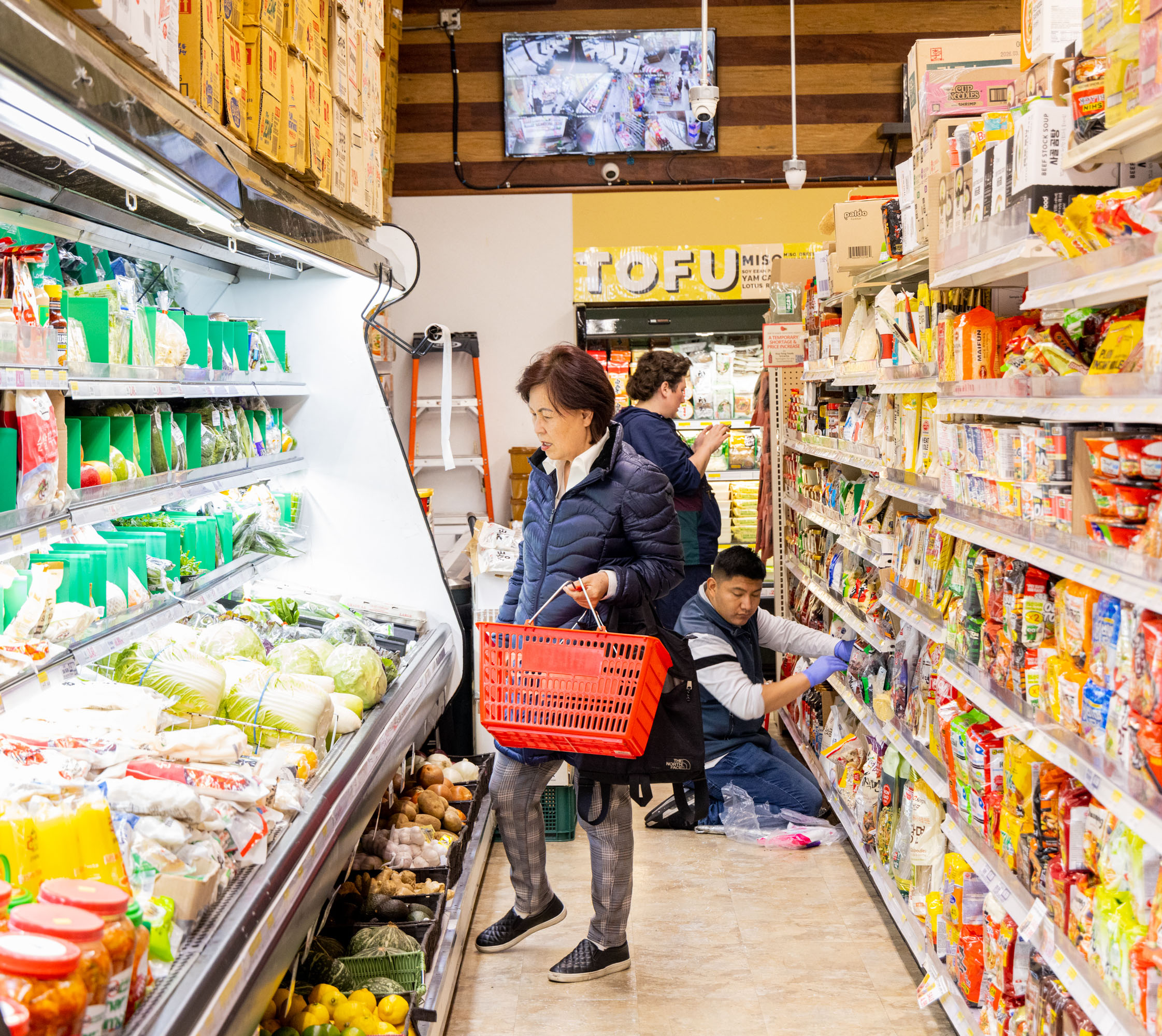A woman with a red basket shops in a grocery aisle. Items are neatly stocked on both sides, and two store employees are organizing products on the right.