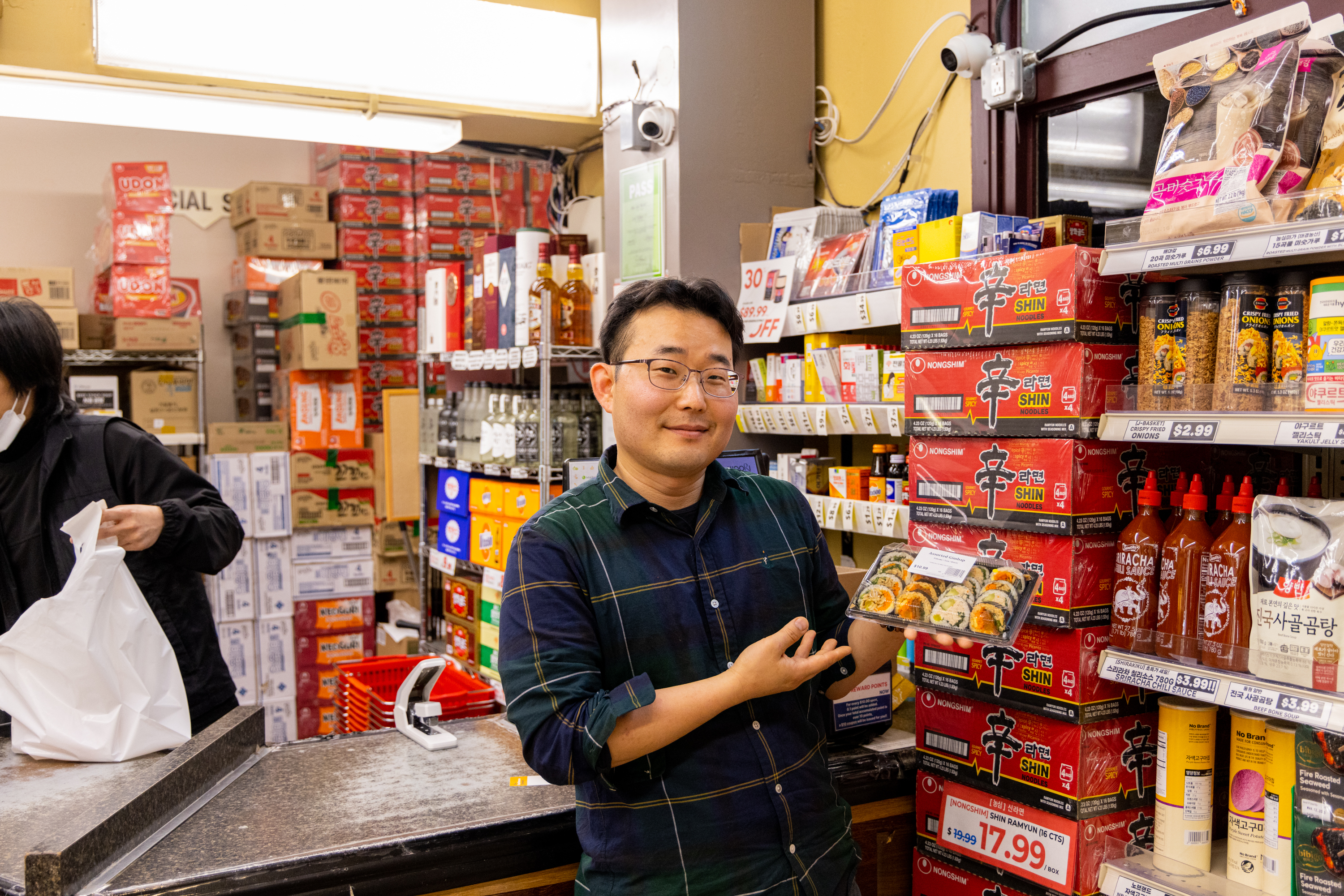 A person in a store smiles while holding a tray of sushi. They stand by shelves stocked with Shin Ramyun noodles, hot sauces, and various groceries. Another person is at the counter.