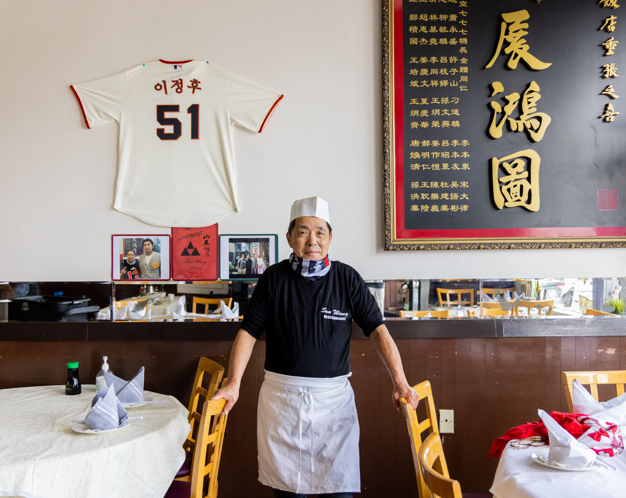 A man dressed as a chef stands in a restaurant beside tables with folded napkins. Behind him are a framed sports jersey, photos, and a large board with text.