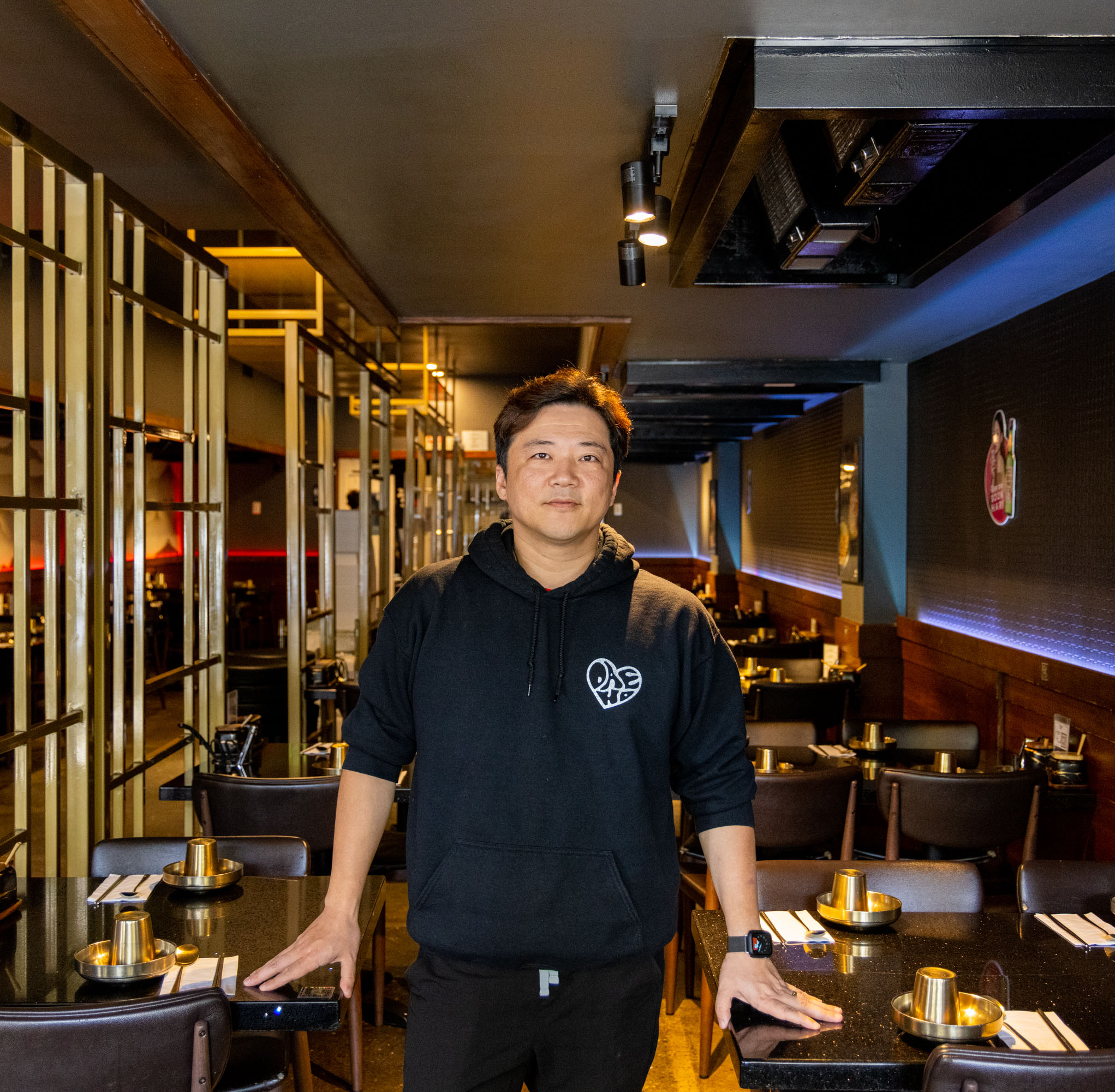A man in a black hoodie stands inside a modern restaurant, with neatly set tables, dim lighting, and contemporary decor featuring metallic and wooden elements.