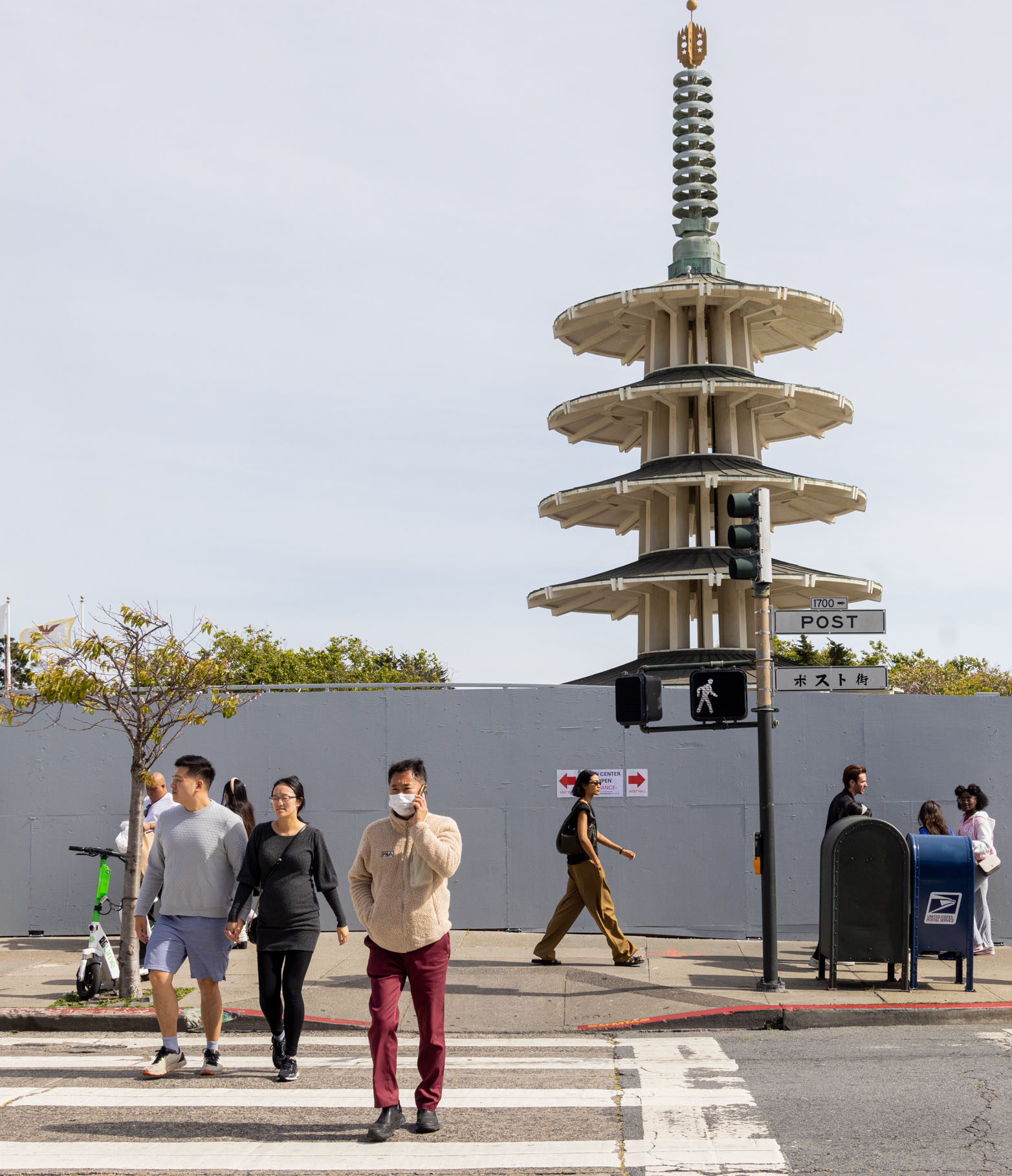 People are crossing a street at a crosswalk with a tall, multi-tiered pagoda structure in the background. There is a traffic light and a street sign reading &quot;Post&quot;.