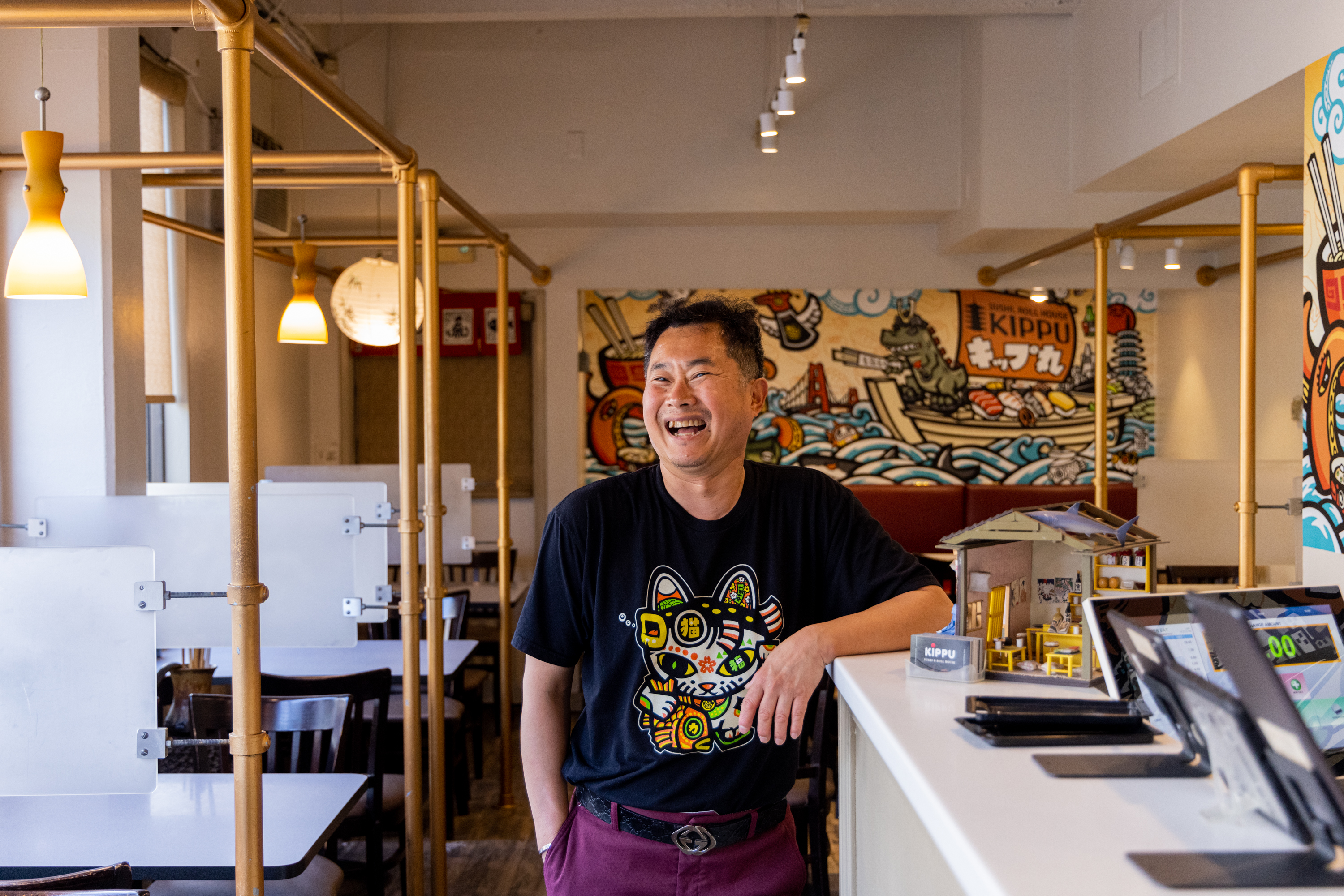 A man is laughing while standing in a brightly lit, colorful restaurant with cartoon-inspired wall art, wearing a black T-shirt and maroon pants.